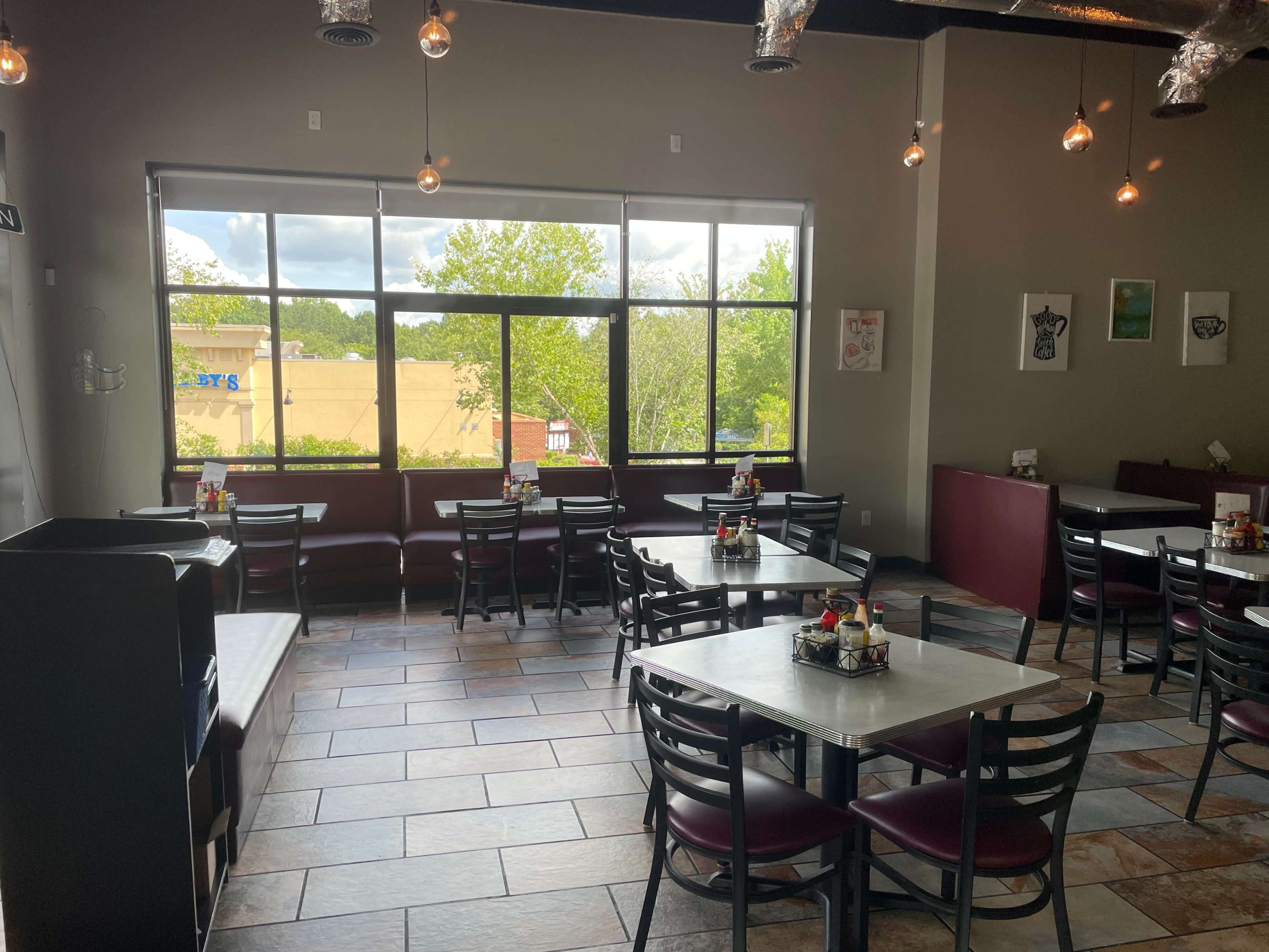 The image shows a spacious restaurant interior with multiple tables and chairs arranged neatly, large windows letting in natural light, and a few decorative elements on the walls.
