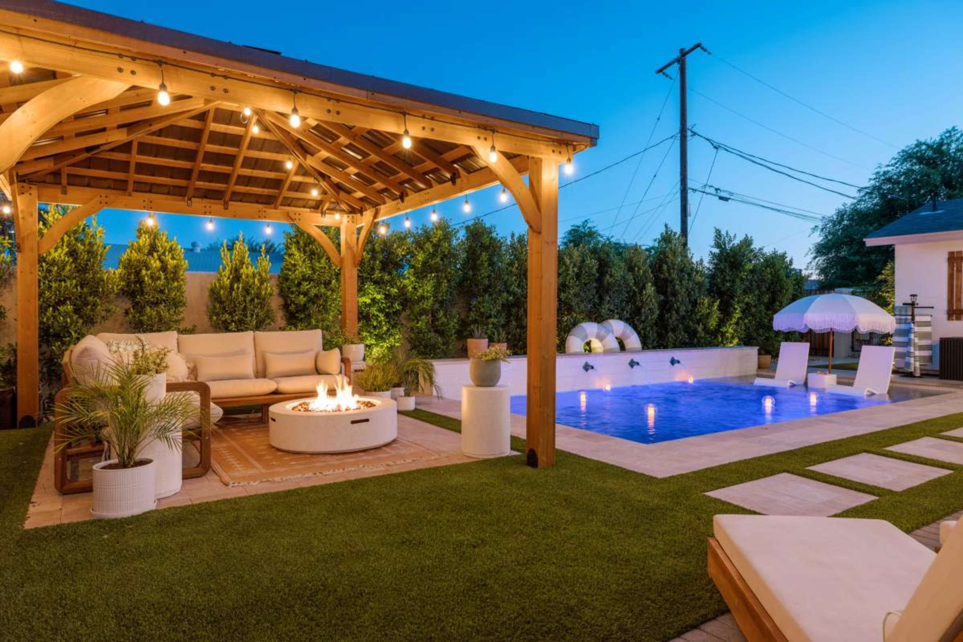 The image shows a landscaped outdoor area featuring a wooden gazebo with seating, a fire pit, a swimming pool, and lounge chairs under a twilight sky.