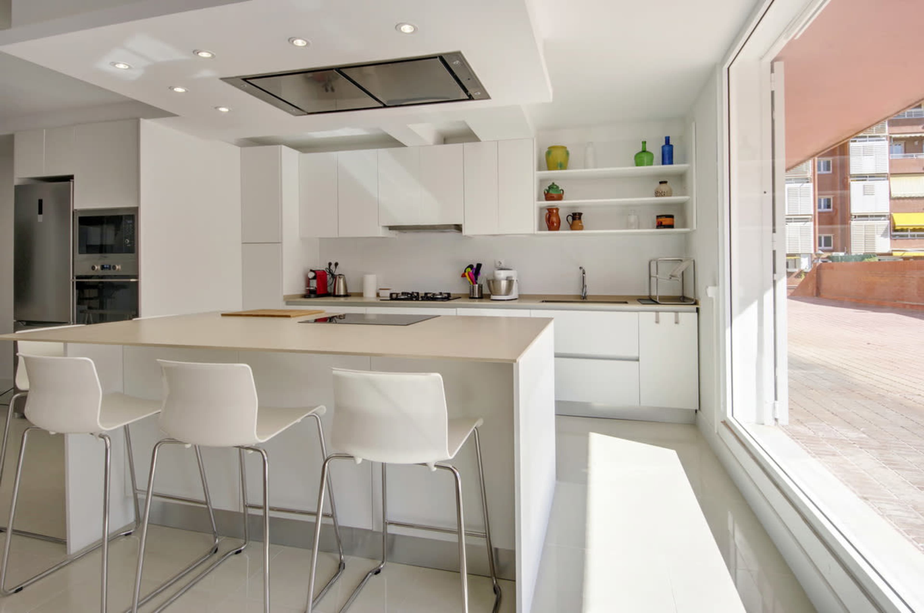 A modern kitchen with white cabinetry, a central island with stools, and large windows allowing natural light to enter the space.