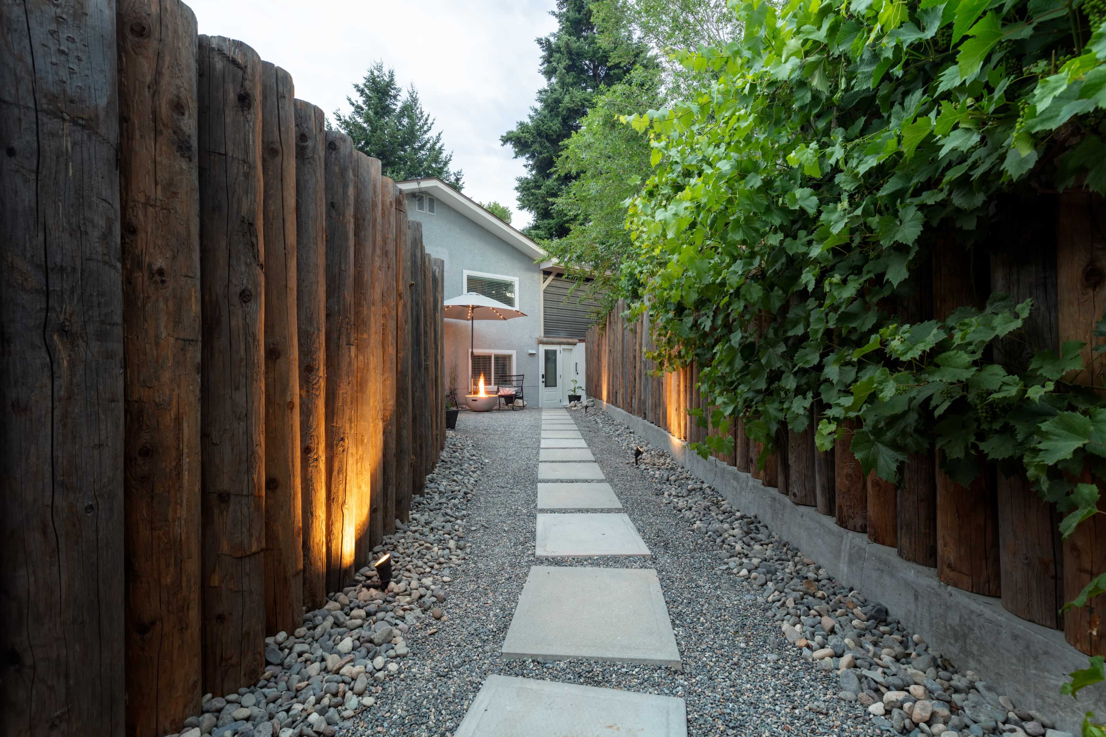 A gravel pathway lined with stepping stones leads to a modern house, enclosed by tall wooden fencing and greenery.