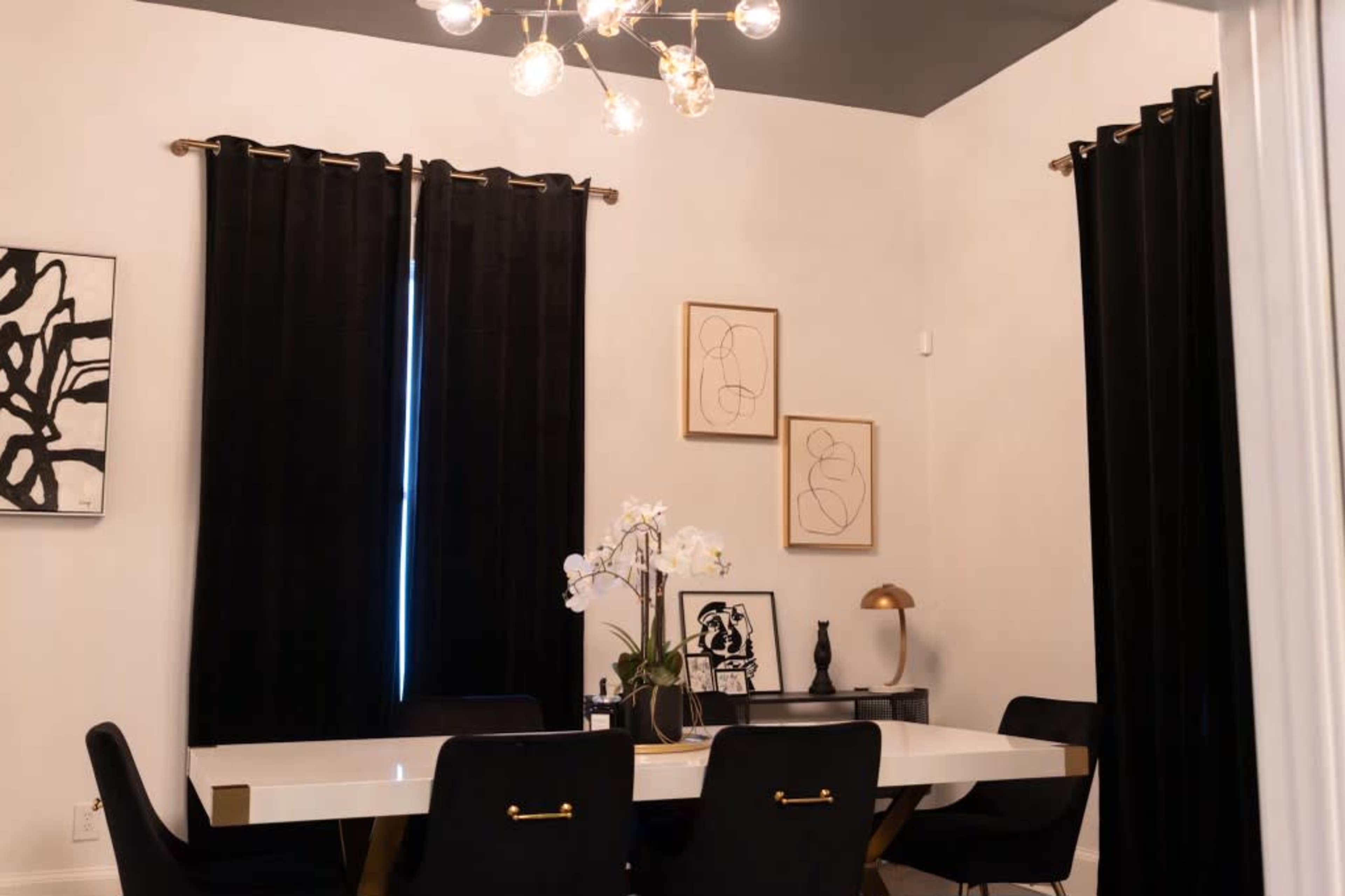 A modern dining area features a white table surrounded by black chairs, with black curtains and framed artwork on the walls.