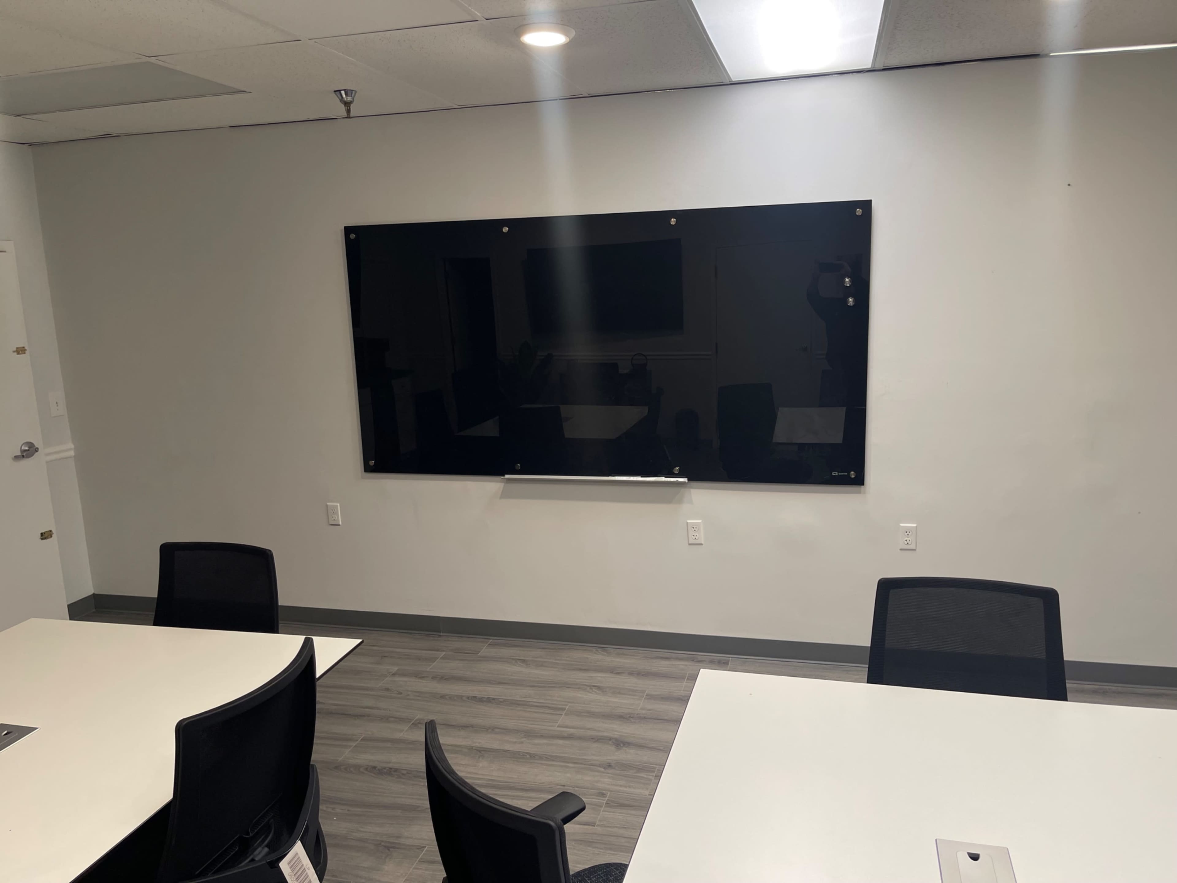 A conference room features a large black presentation screen mounted on a gray wall, with several tables and chairs arranged below it.