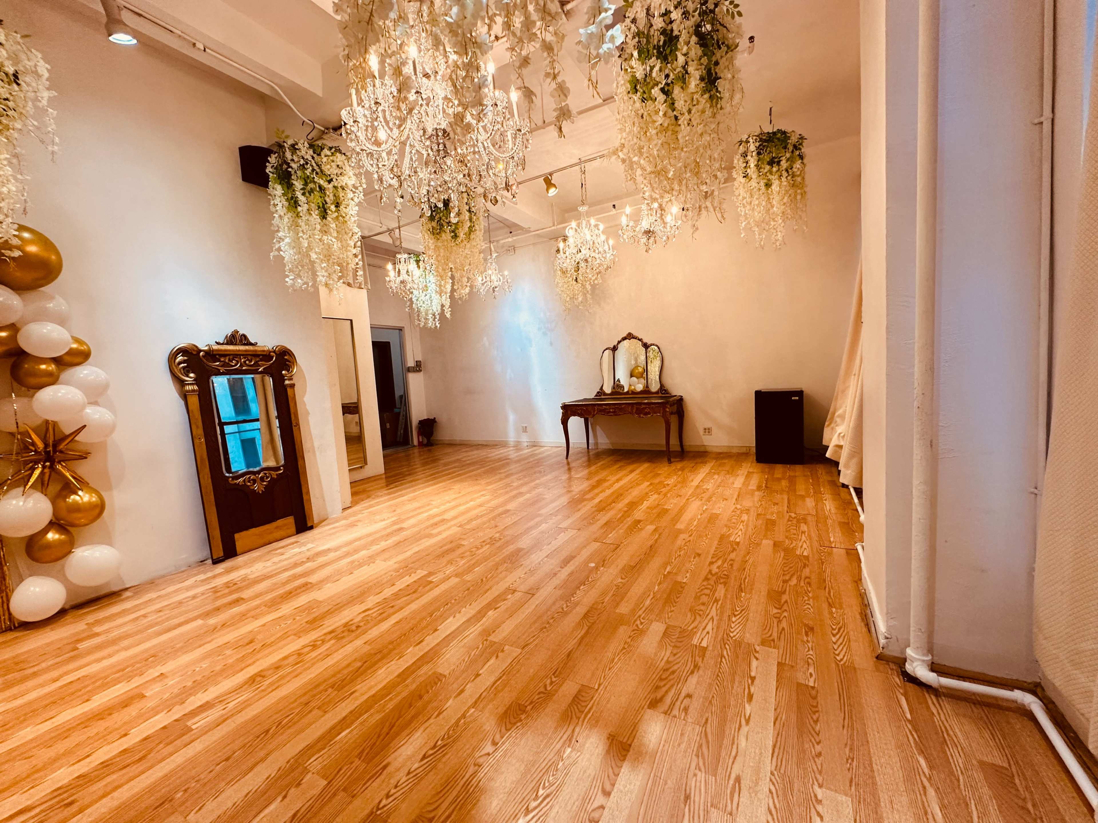 A spacious room with wooden flooring, adorned with chandeliers and hanging greenery, featuring a large mirror and a vintage-style table against one wall.