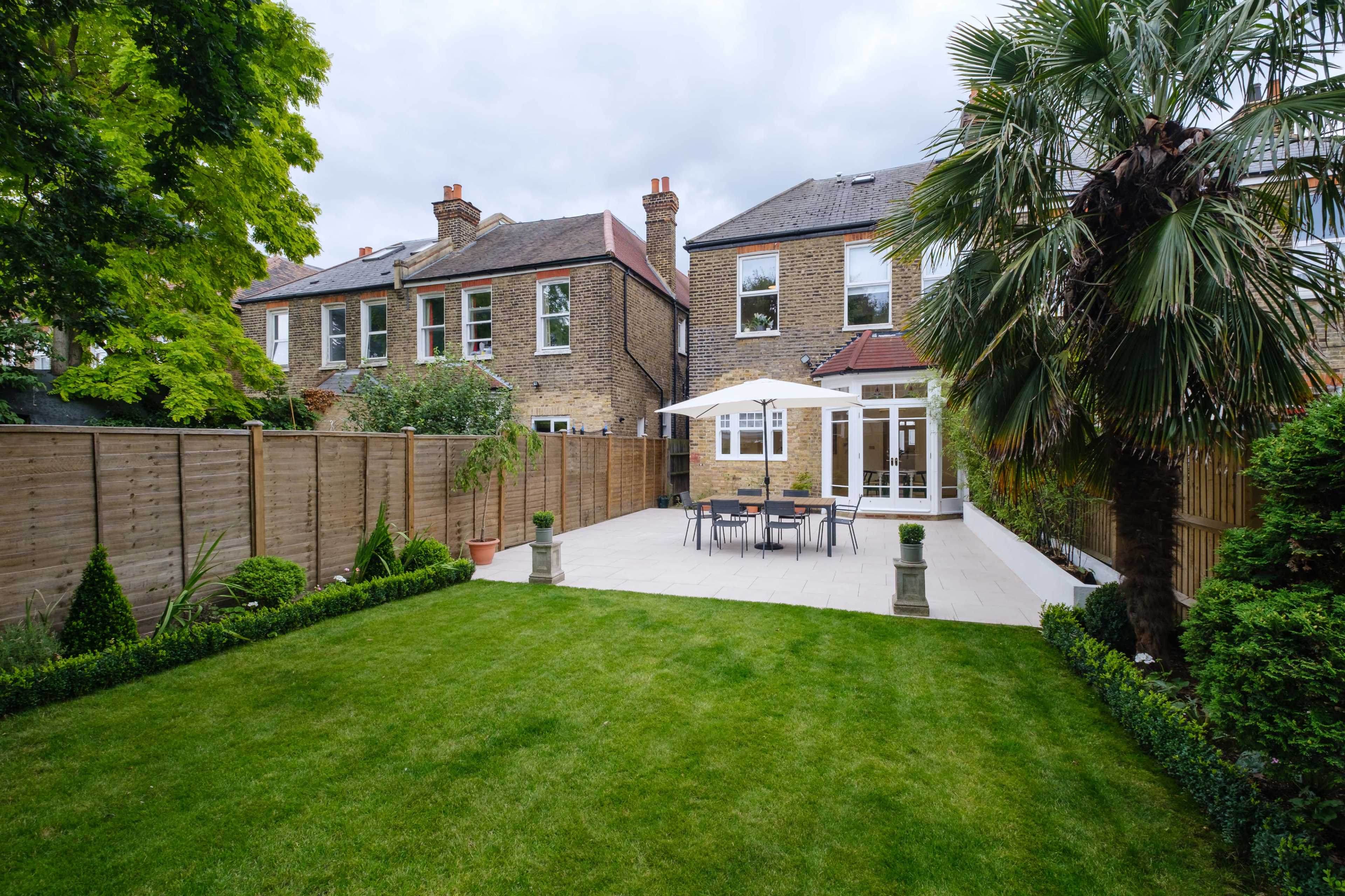 A backyard featuring a small patio set under a white umbrella, surrounded by well-maintained grass and landscaping, with two houses and a wooden fence in the background.