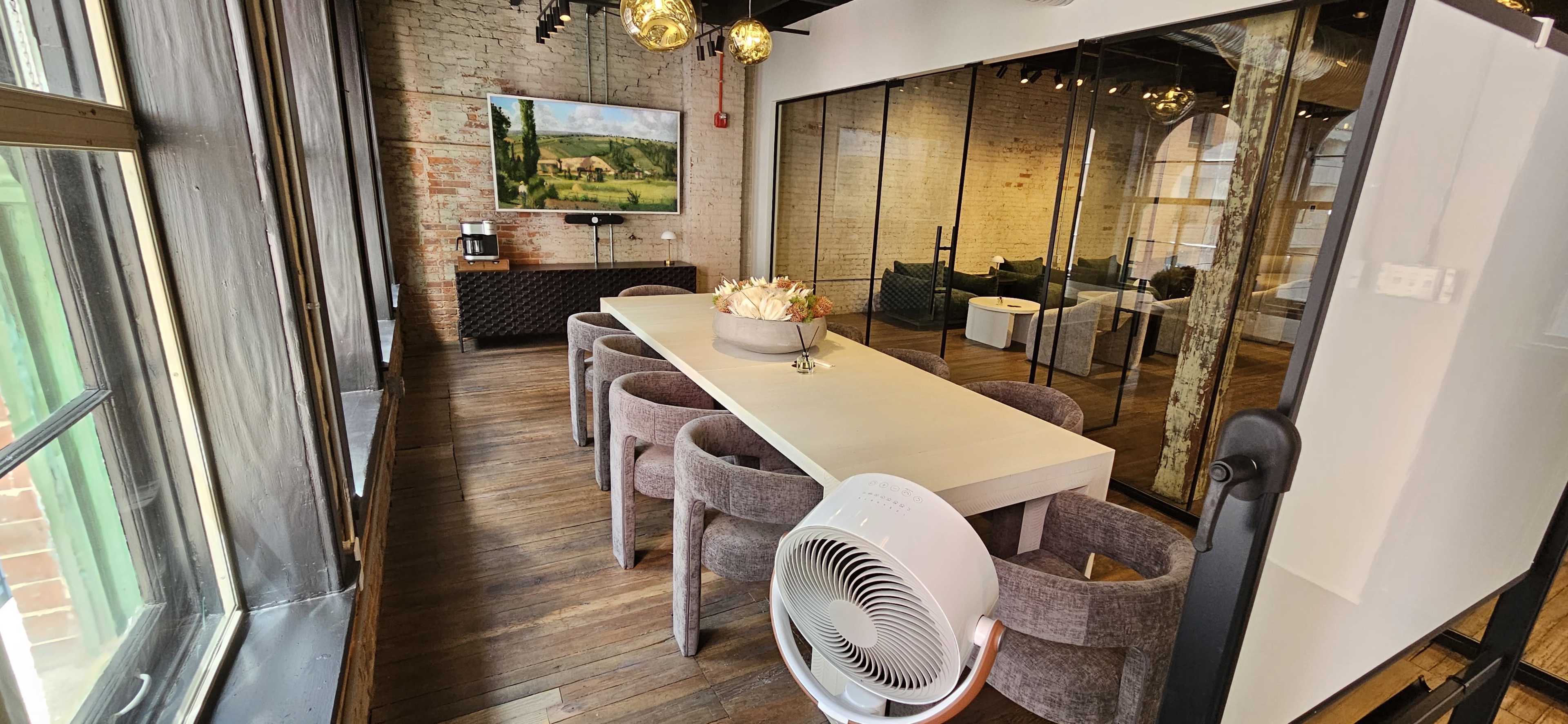 A long white table is surrounded by gray chairs in a well-lit meeting room featuring exposed brick walls and large windows.
