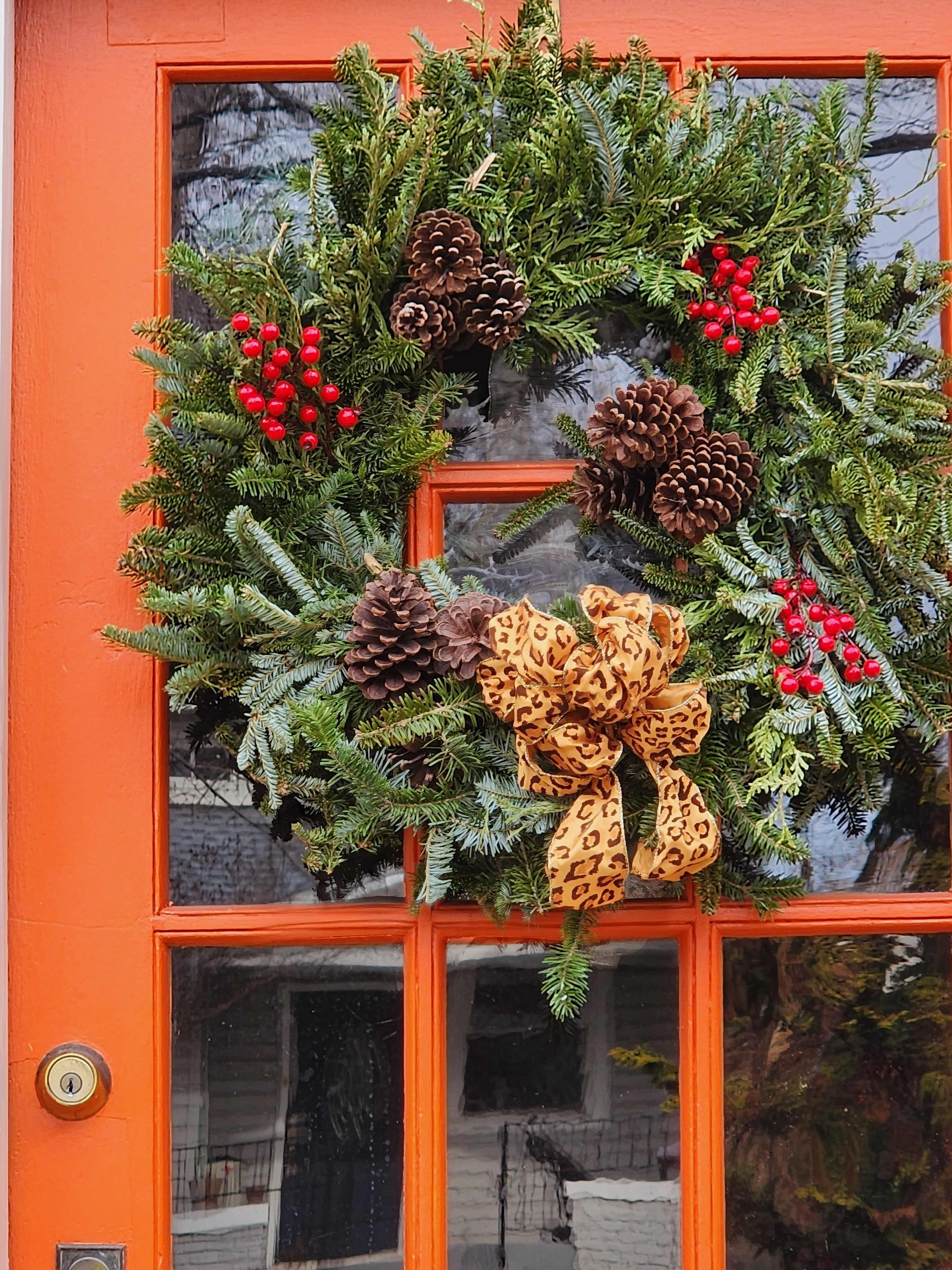 A green wreath adorned with pinecones, red berries, and a leopard print bow is hung on an orange door.