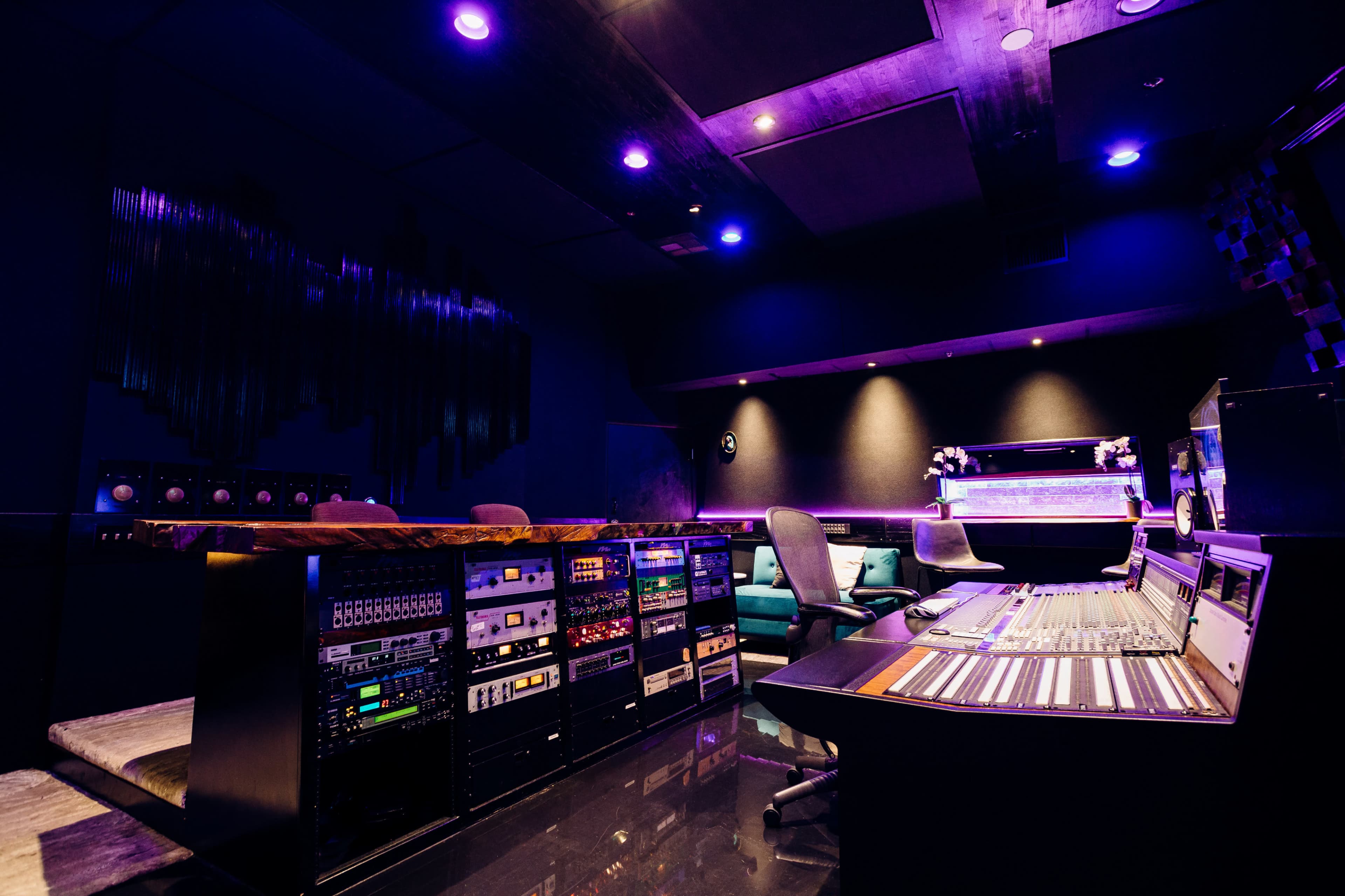 A modern recording studio with various audio equipment and a large mixing console under purple lighting.