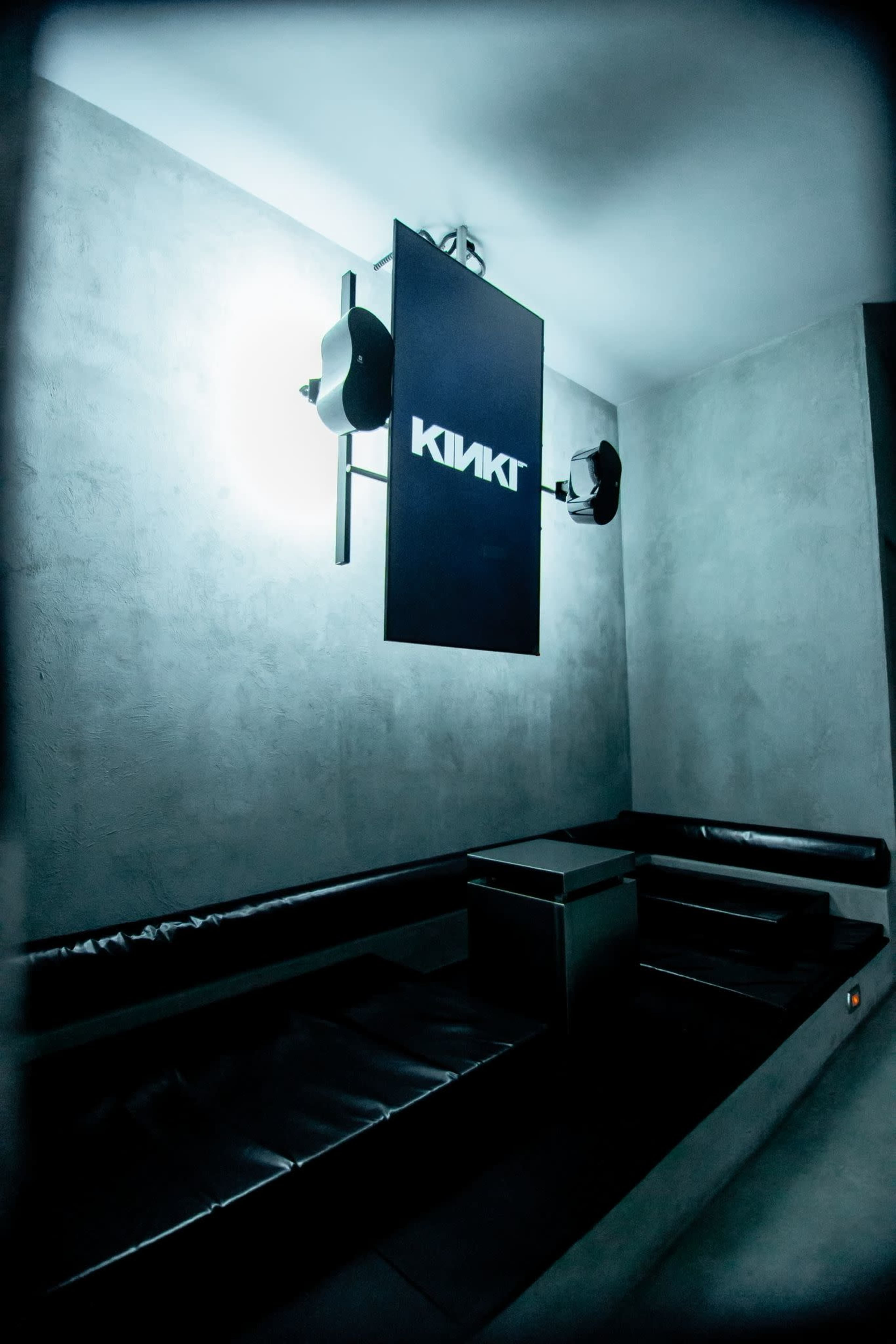 The image shows a minimalist, modern room with gray walls and a sleek, black bench, featuring a sign that reads "KINKT" illuminated by overhead lights.