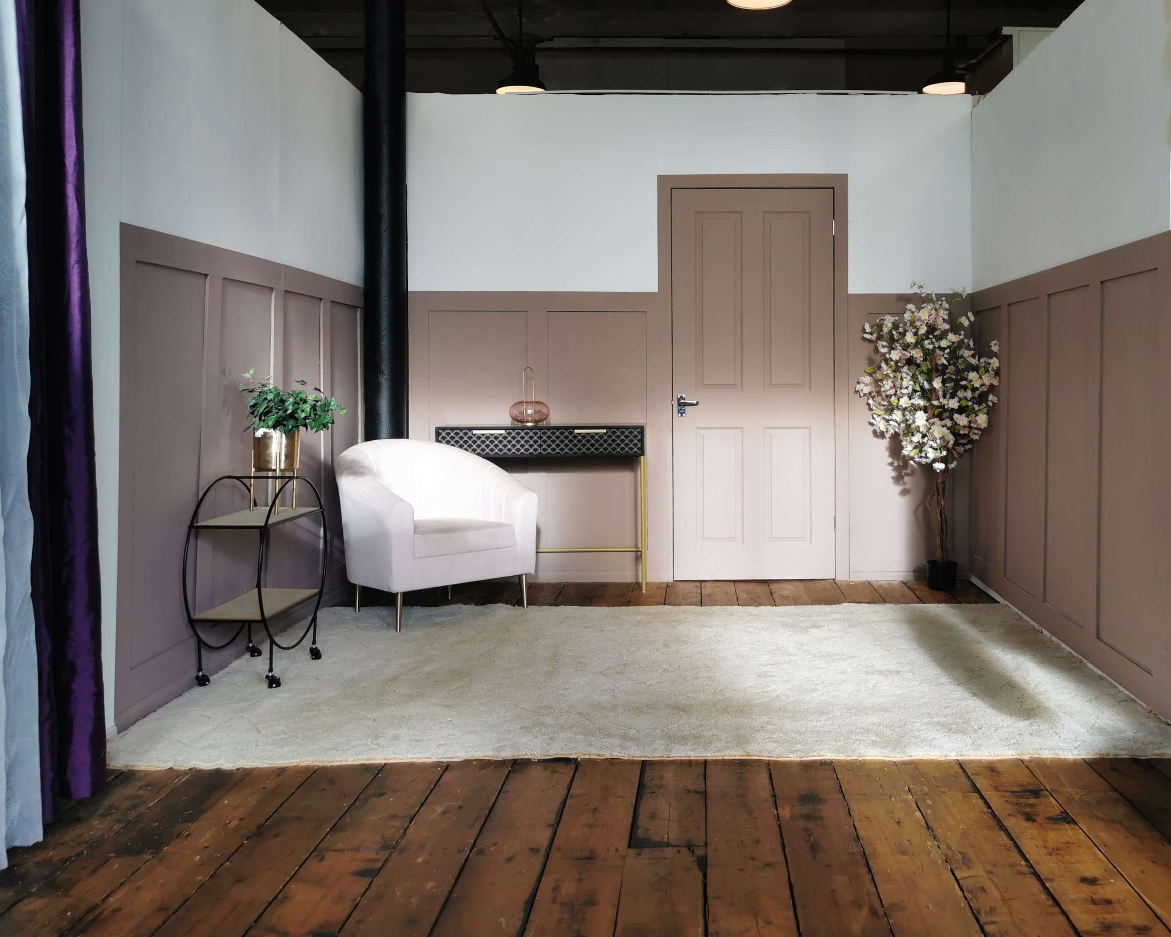 The image shows a room with a light-colored wooden floor, a pink armchair, a small circular table, a decorative flower arrangement, and a closed door against a neutral-colored wall.