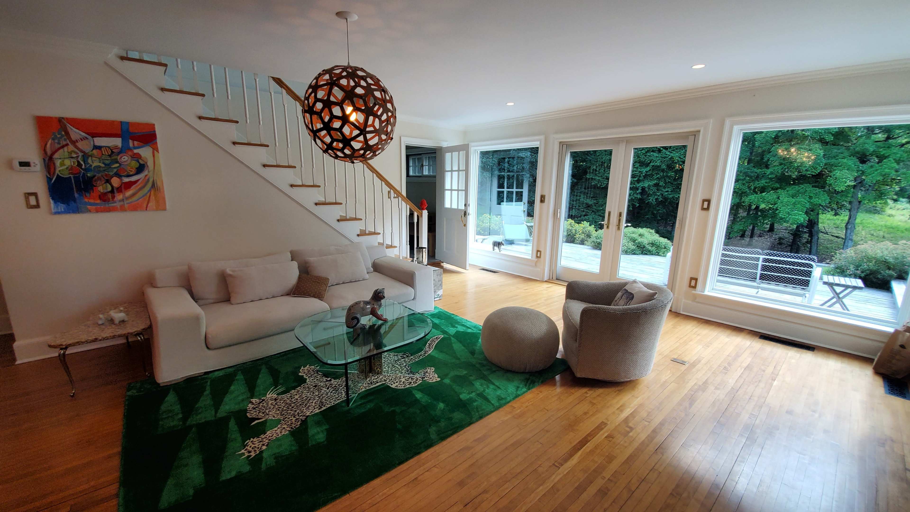 A living room features a white sofa, a circular glass coffee table, a green rug with a leopard print design, and large windows overlooking a patio.