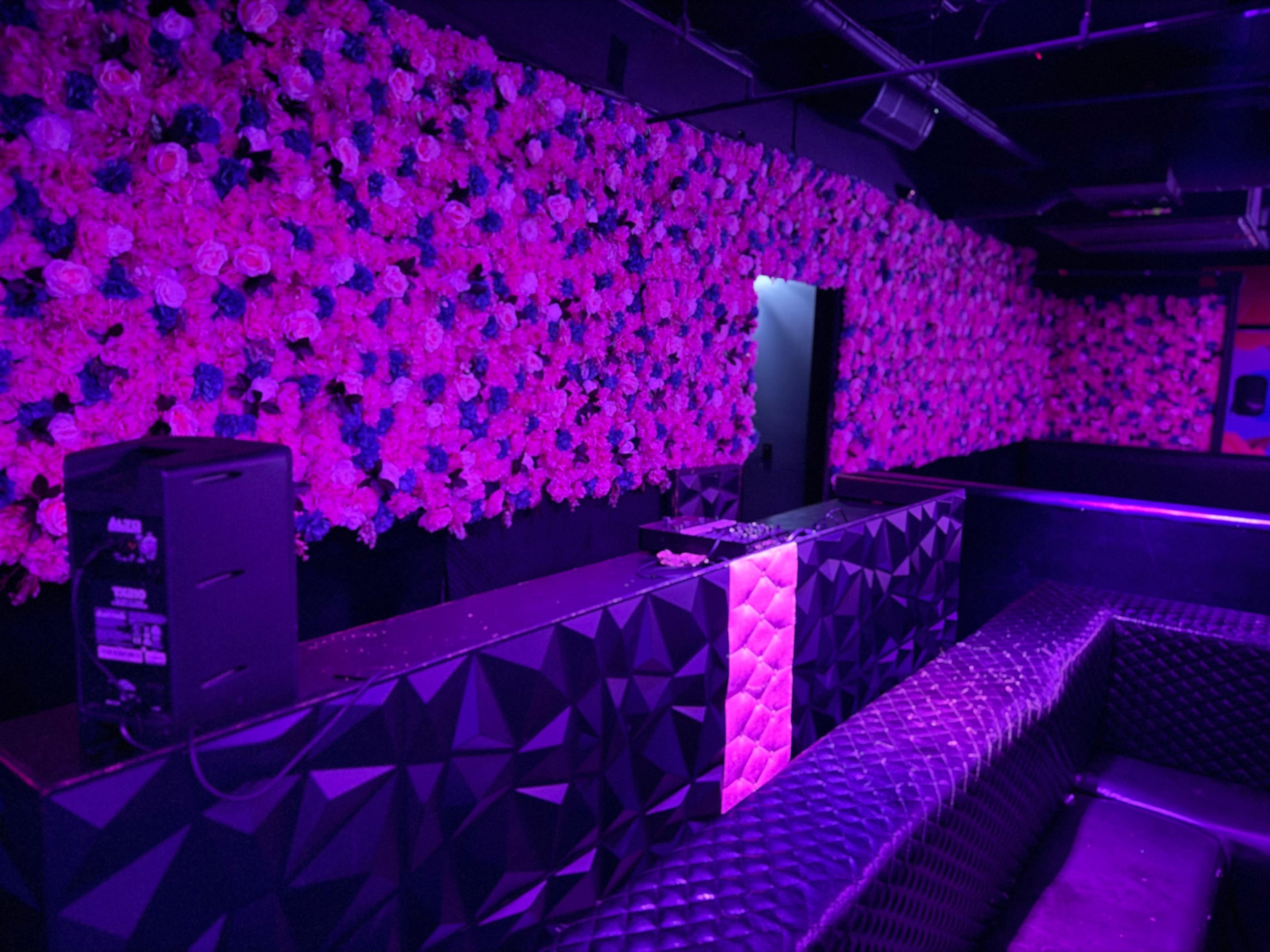 The image shows a nightlife venue featuring a wall covered in artificial flowers, with a dark, geometric bar and seating illuminated by purple lighting.