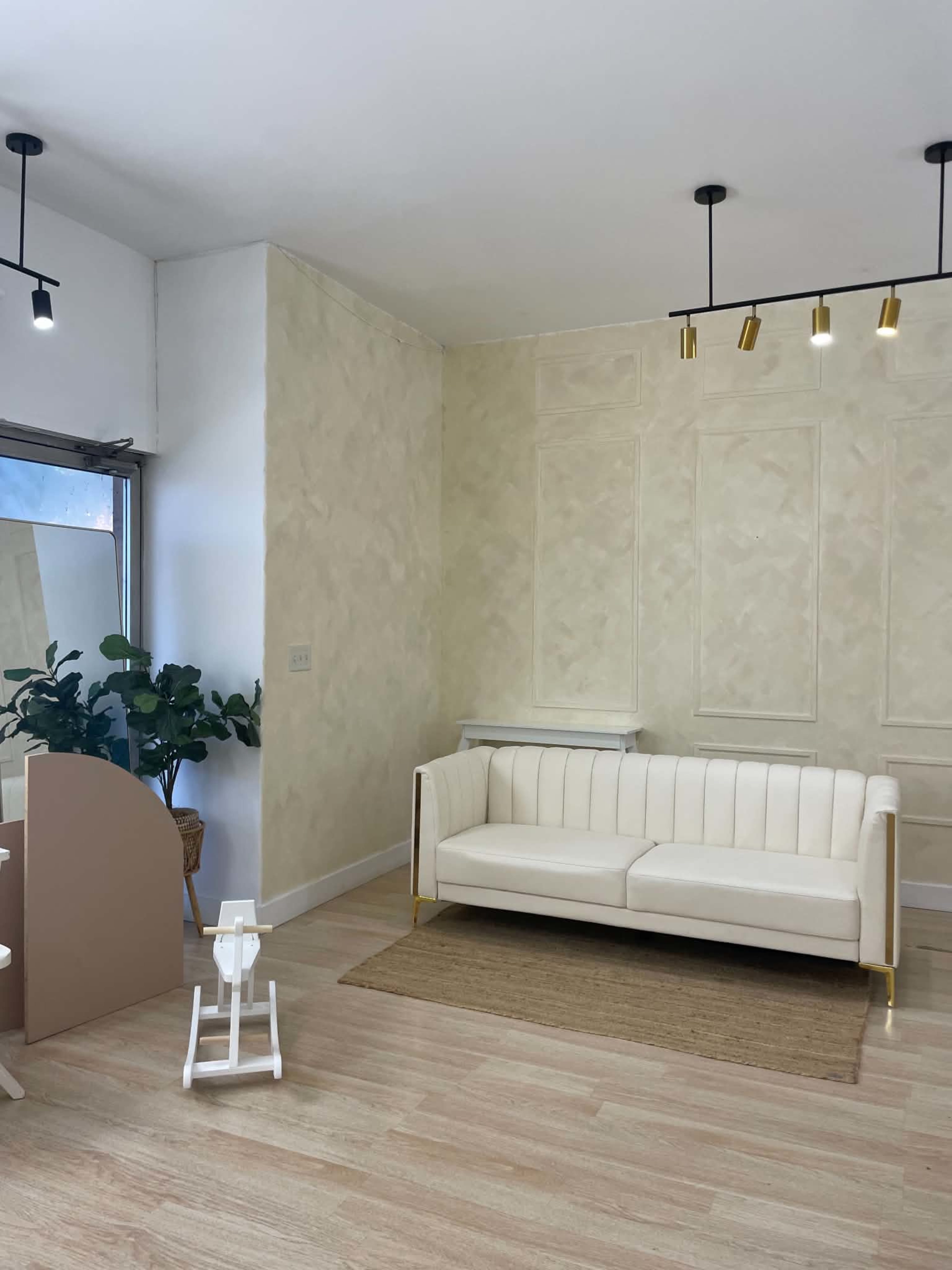 The image shows a minimalist room featuring a white sofa, a small rug, a plant, and a softly textured wall with pendant lighting.