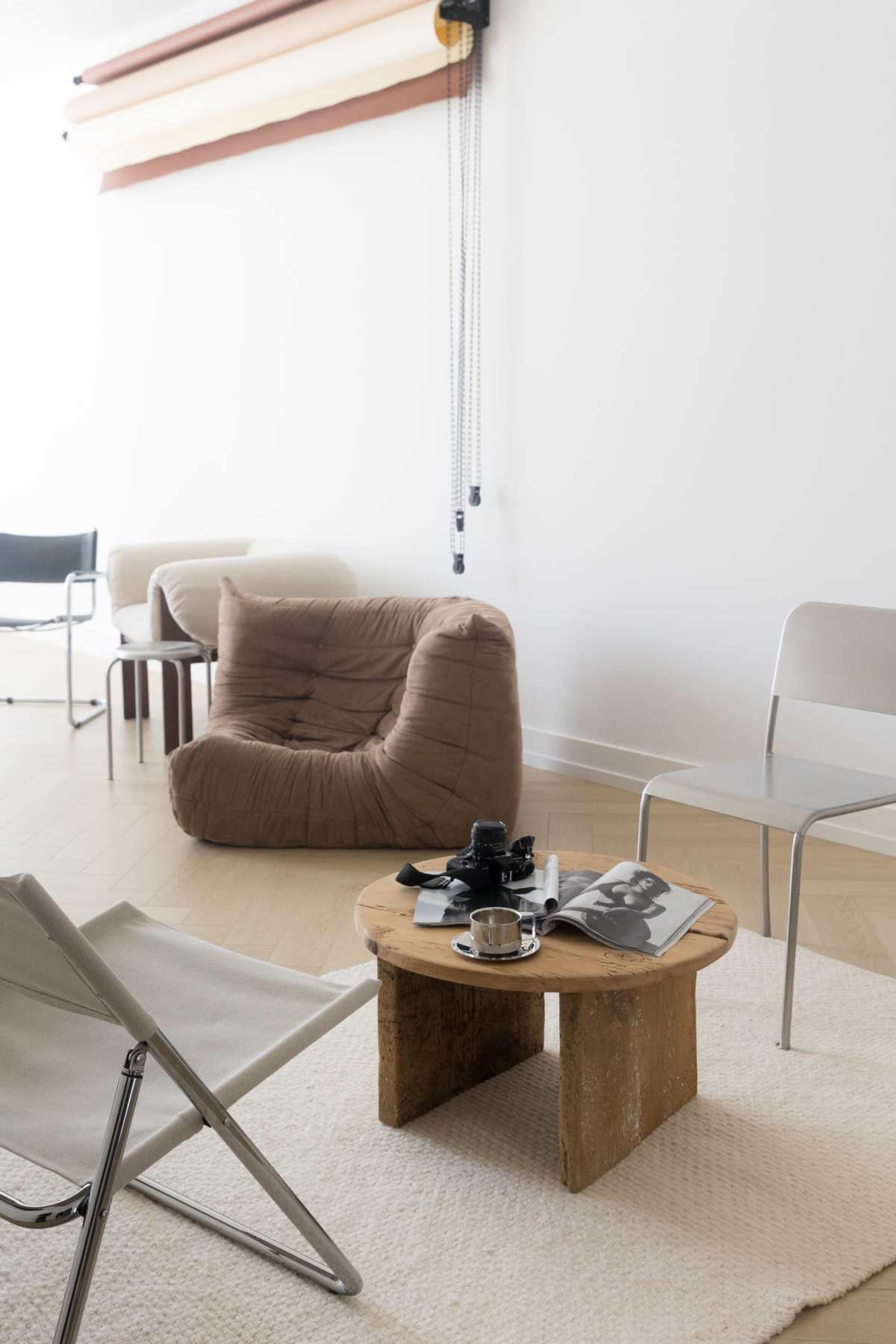 The image depicts a minimalist living room with a wooden coffee table, a stylish magazine, a coffee cup, and various seating options, including a bean bag and metal chairs.