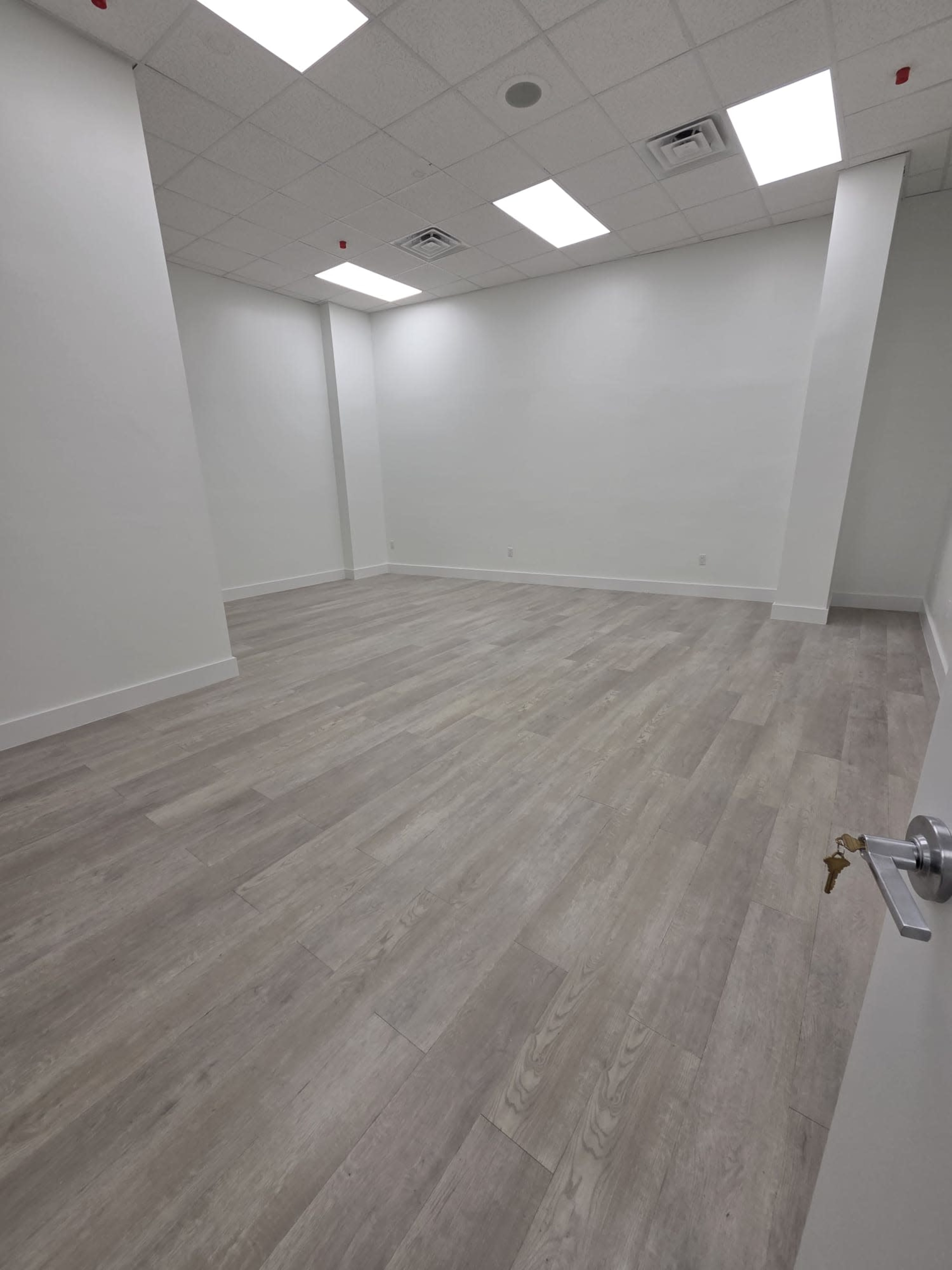 The image shows an empty room with light gray wooden flooring and plain white walls, illuminated by overhead fluorescent lights.