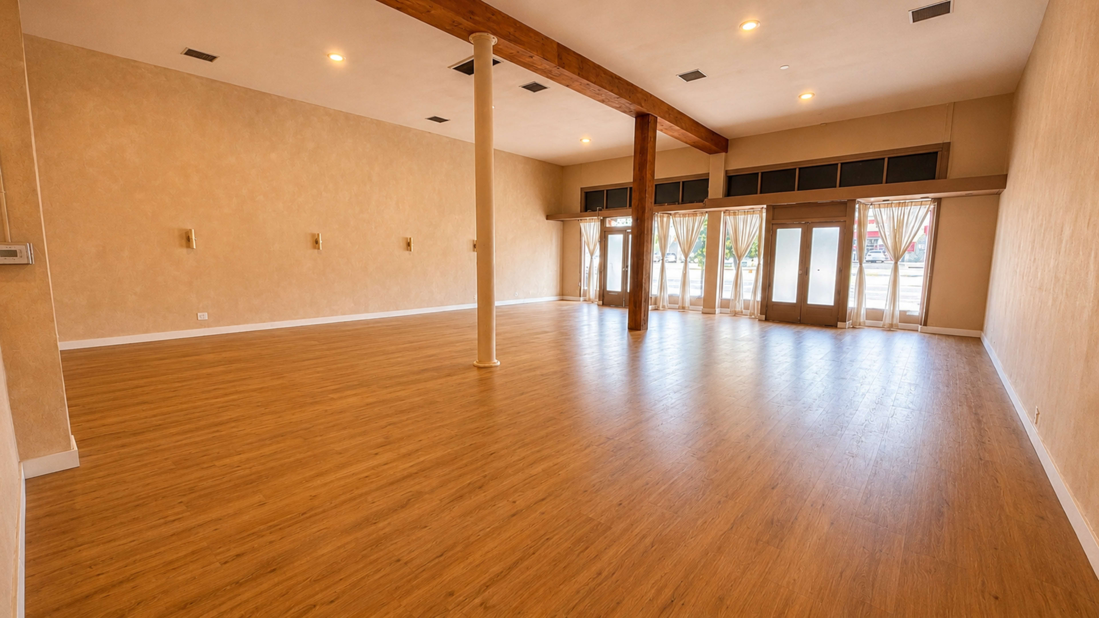 The image shows an empty room with wooden flooring, large windows draped with curtains, and a high ceiling supported by exposed beams and a column.