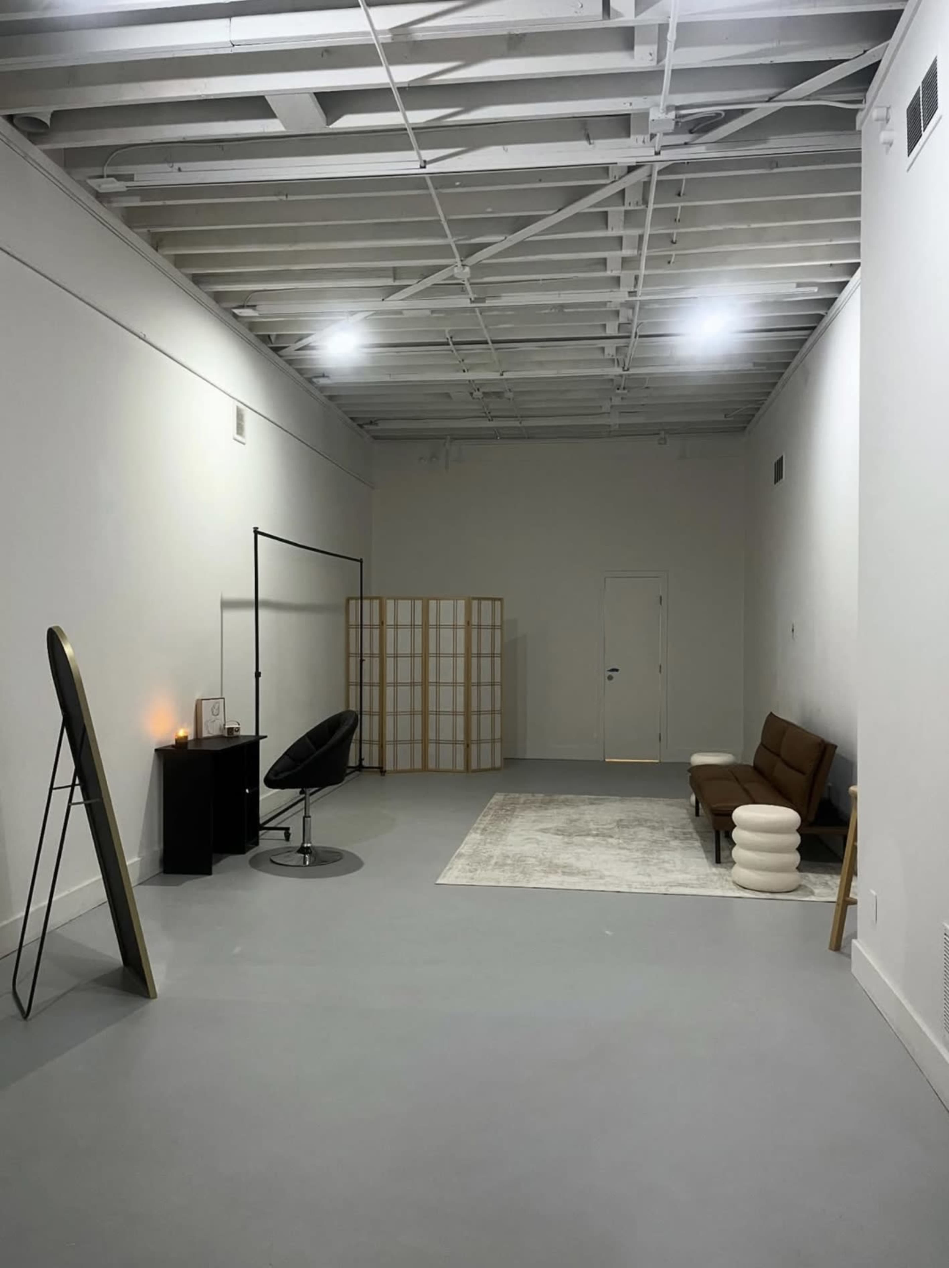 The image shows a spacious, minimalistic room with a gray floor, a wooden couch, a large mirror, a black chair, a small table, and a room divider.