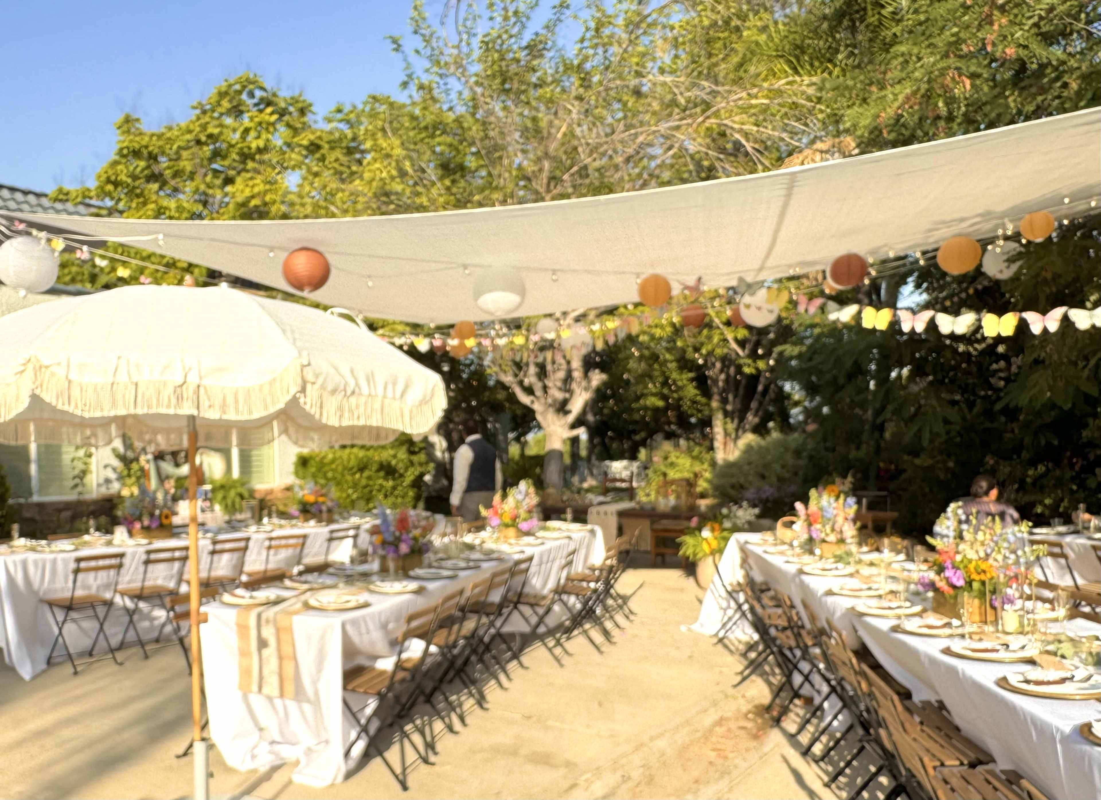 A dining setup for an outdoor event features long tables adorned with flowers, tableware, and decorative string lights under fabric canopies.