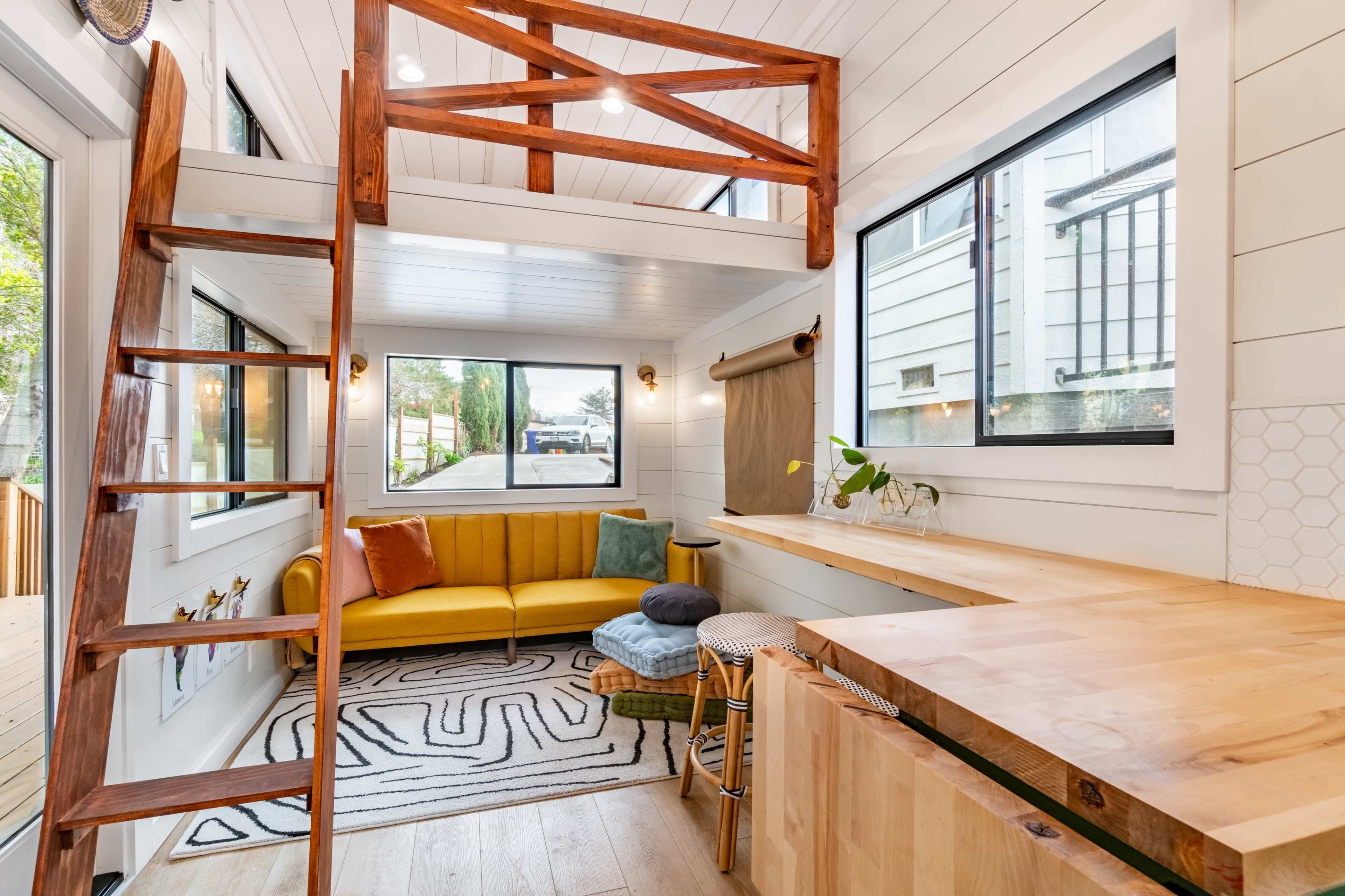 A bright, modern interior of a small living space featuring a yellow sofa, a wooden ladder leading to a loft area, and a small kitchen bar with stools.