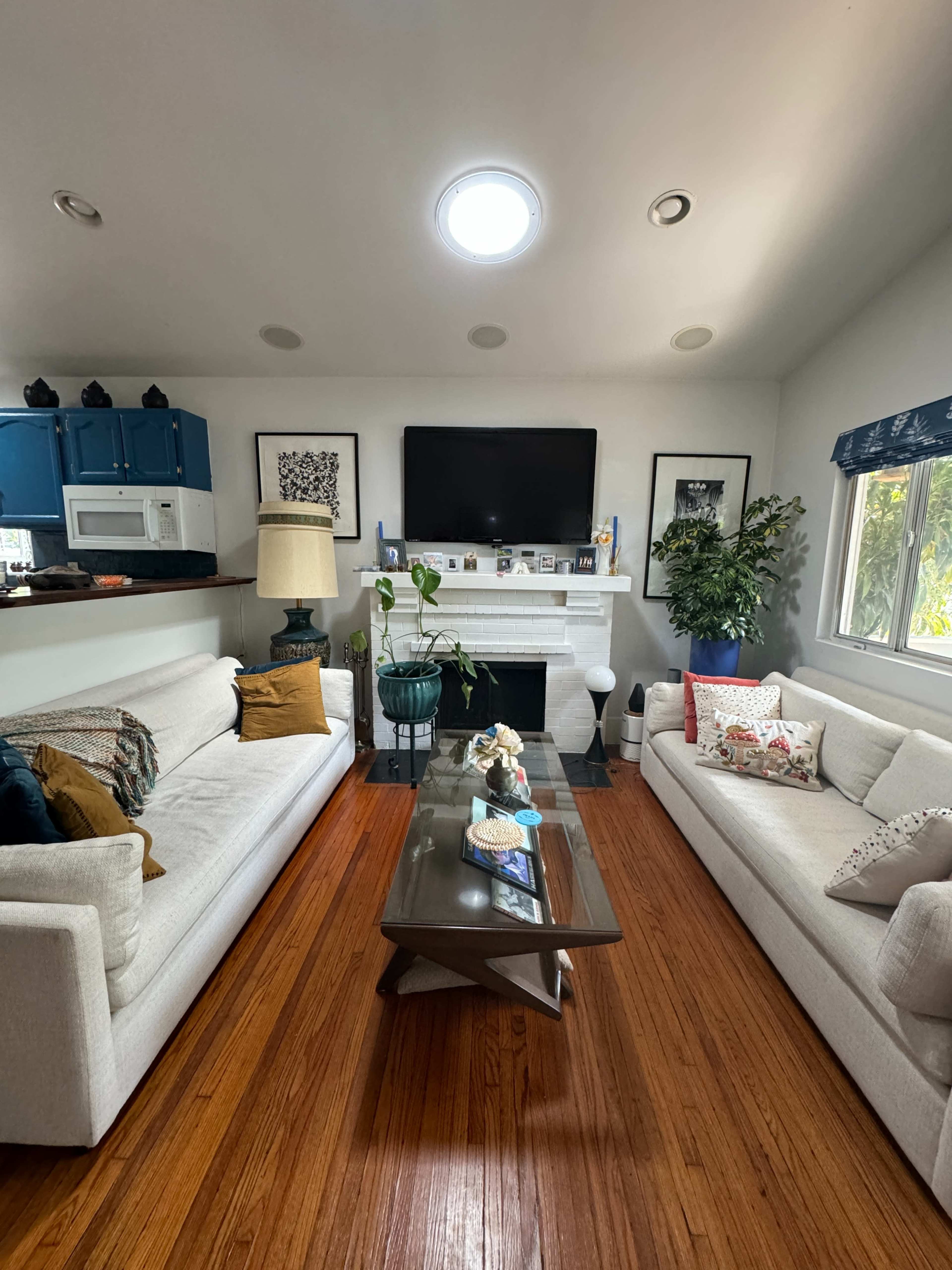The living room features two white sofas, a wooden coffee table, and a television mounted above a fireplace, with decorative plants and artwork on the walls.