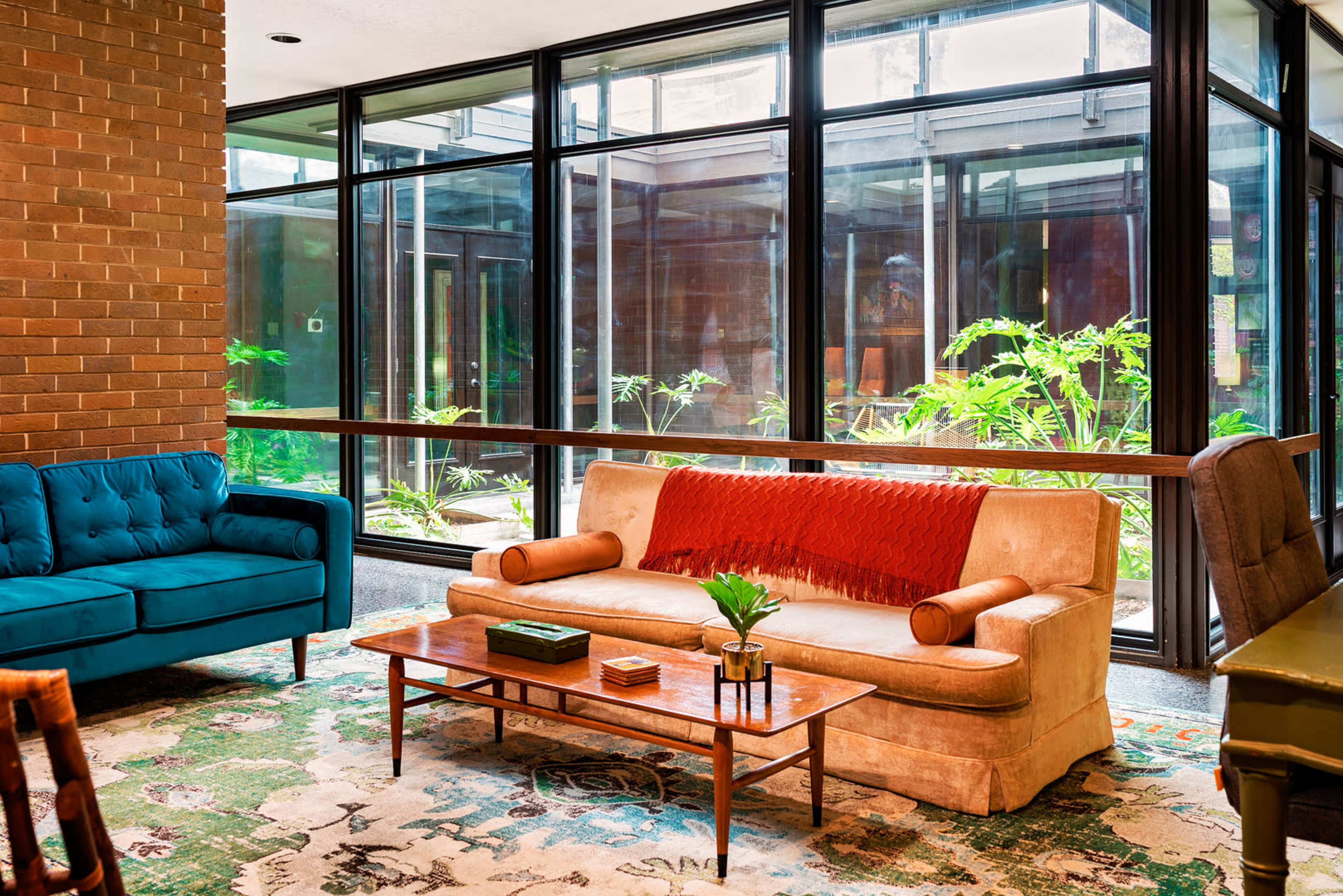 The image depicts a modern living space featuring a beige sofa with round armrests, a wooden coffee table, and large windows that allow natural light to illuminate indoor plants.