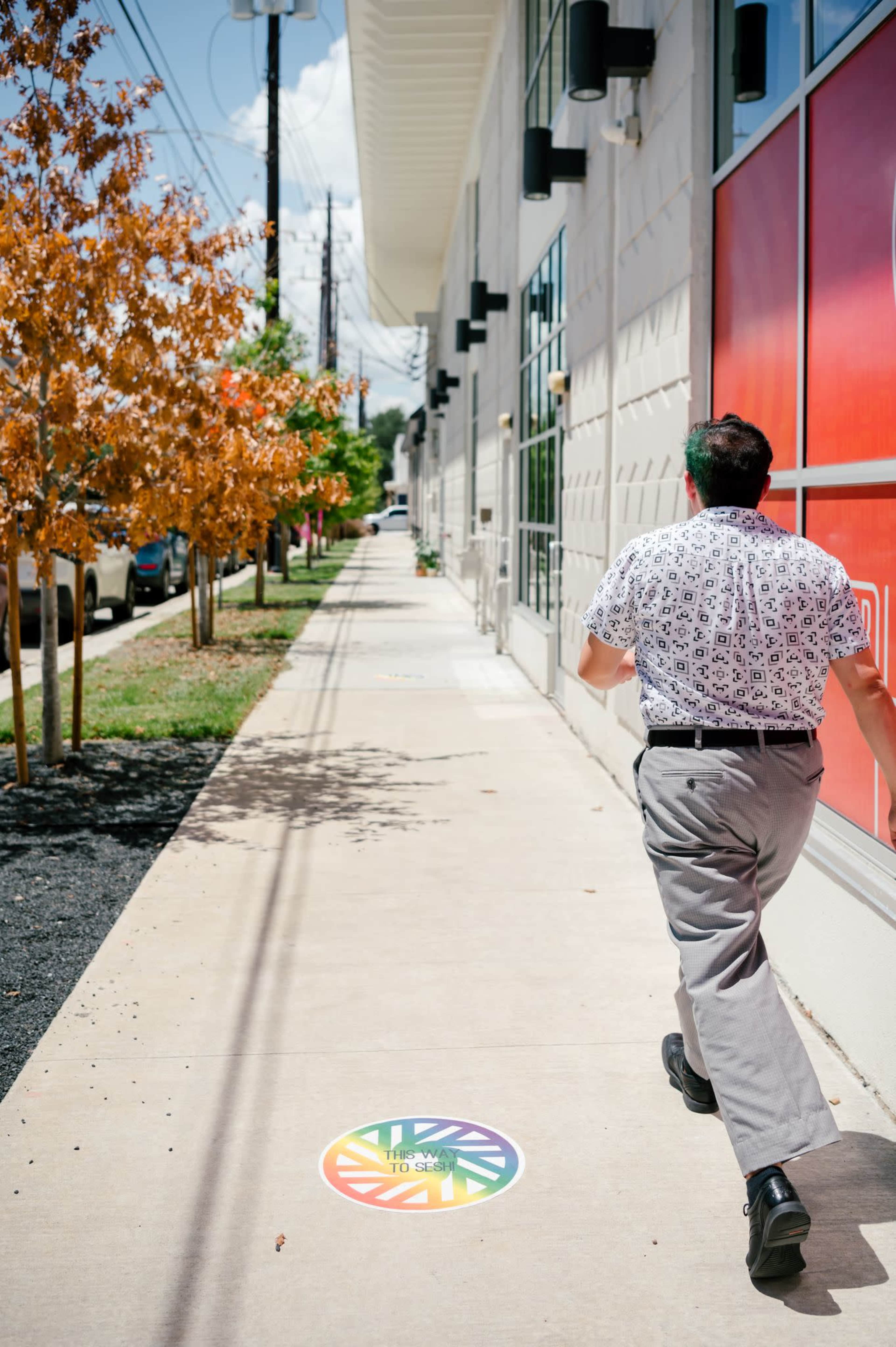 A man walks along a sidewalk beside a building, with colorful trees lining the street and a circular design on the ground.