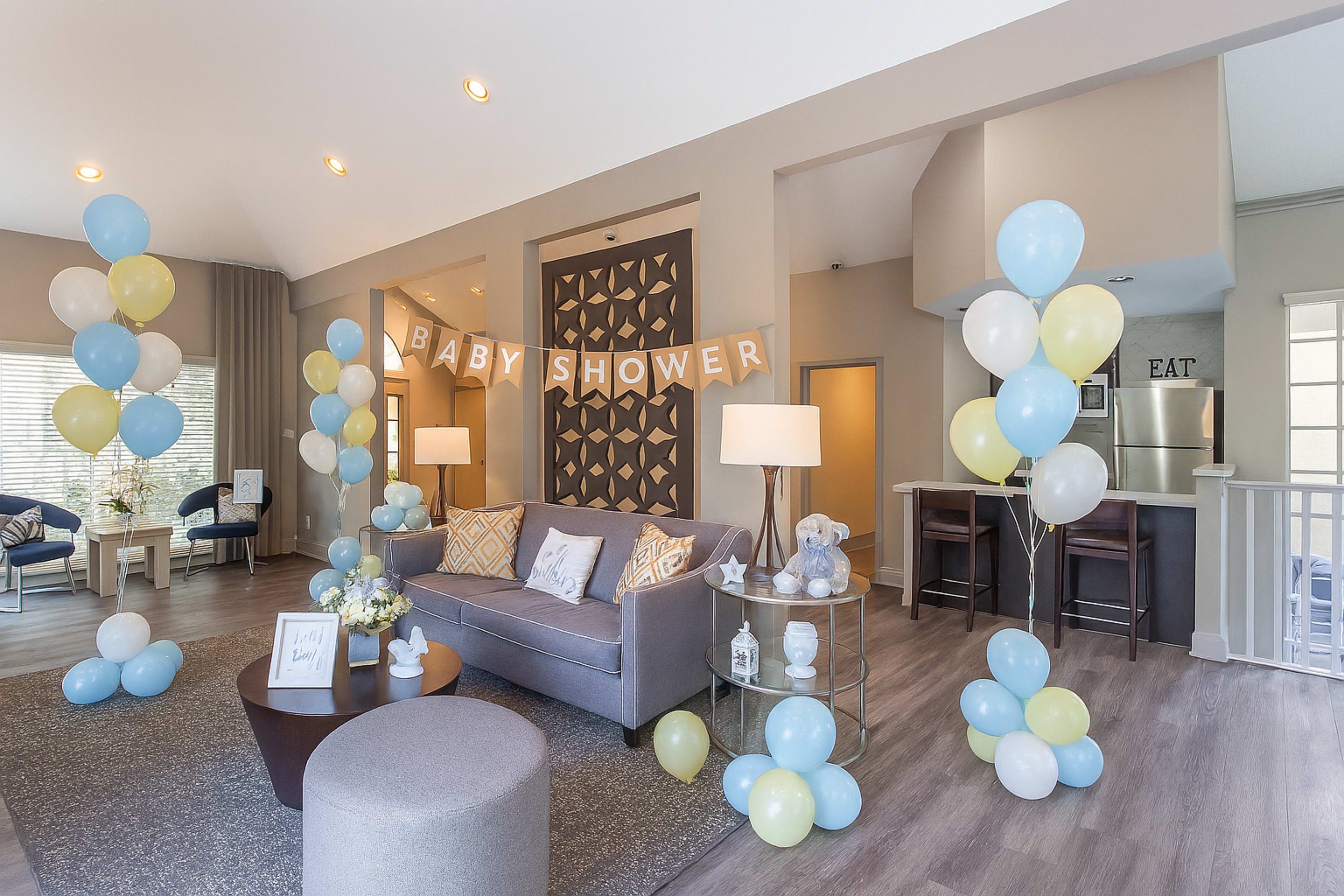 The image shows a decorated living area for a baby shower, featuring balloons in light blue and yellow, a banner that says "BABY SHOWER," and a cozy seating arrangement.