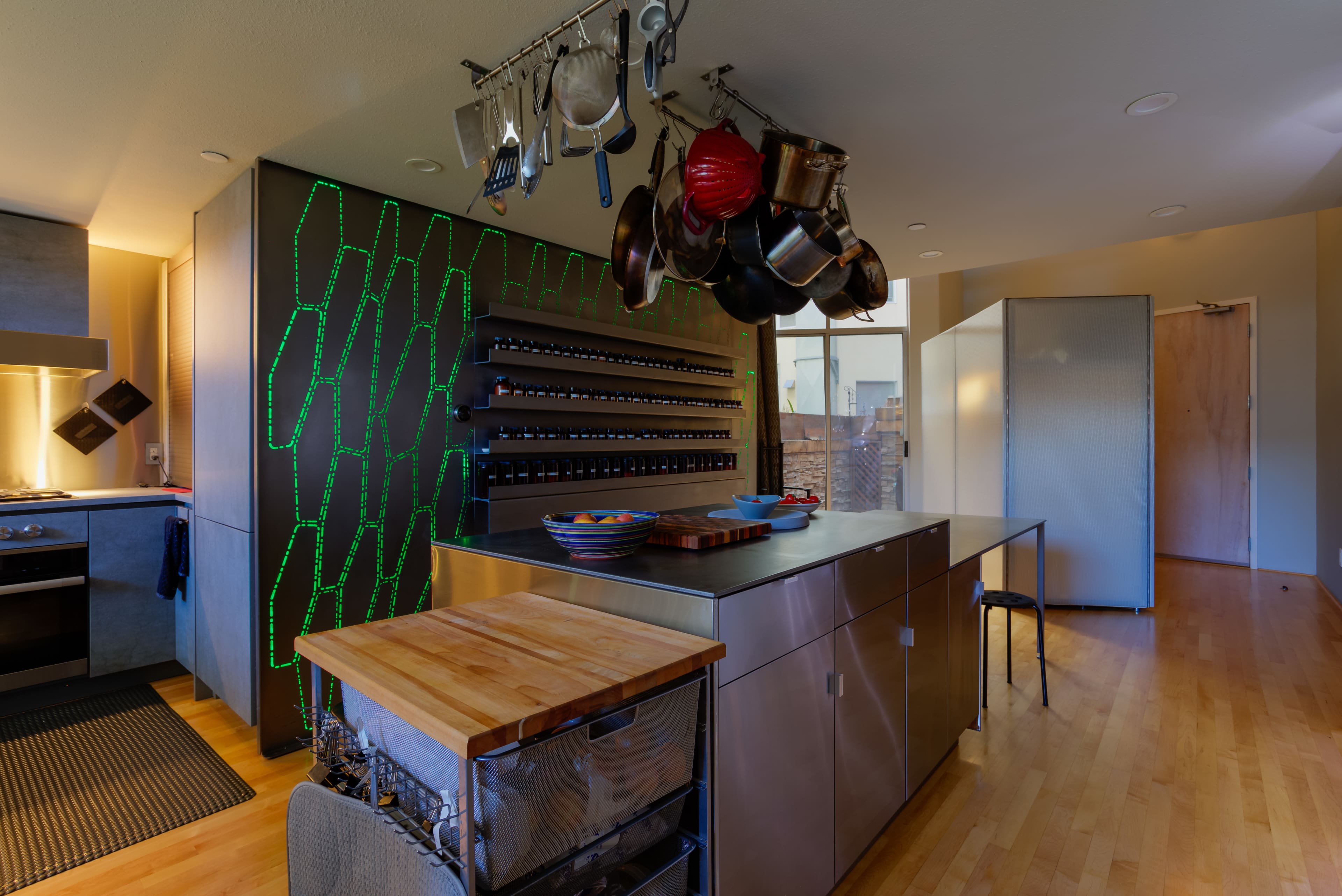 A modern kitchen featuring a central island with a wooden countertop, metal cabinetry, hanging pots, and a wall of wine storage with illuminated accents.