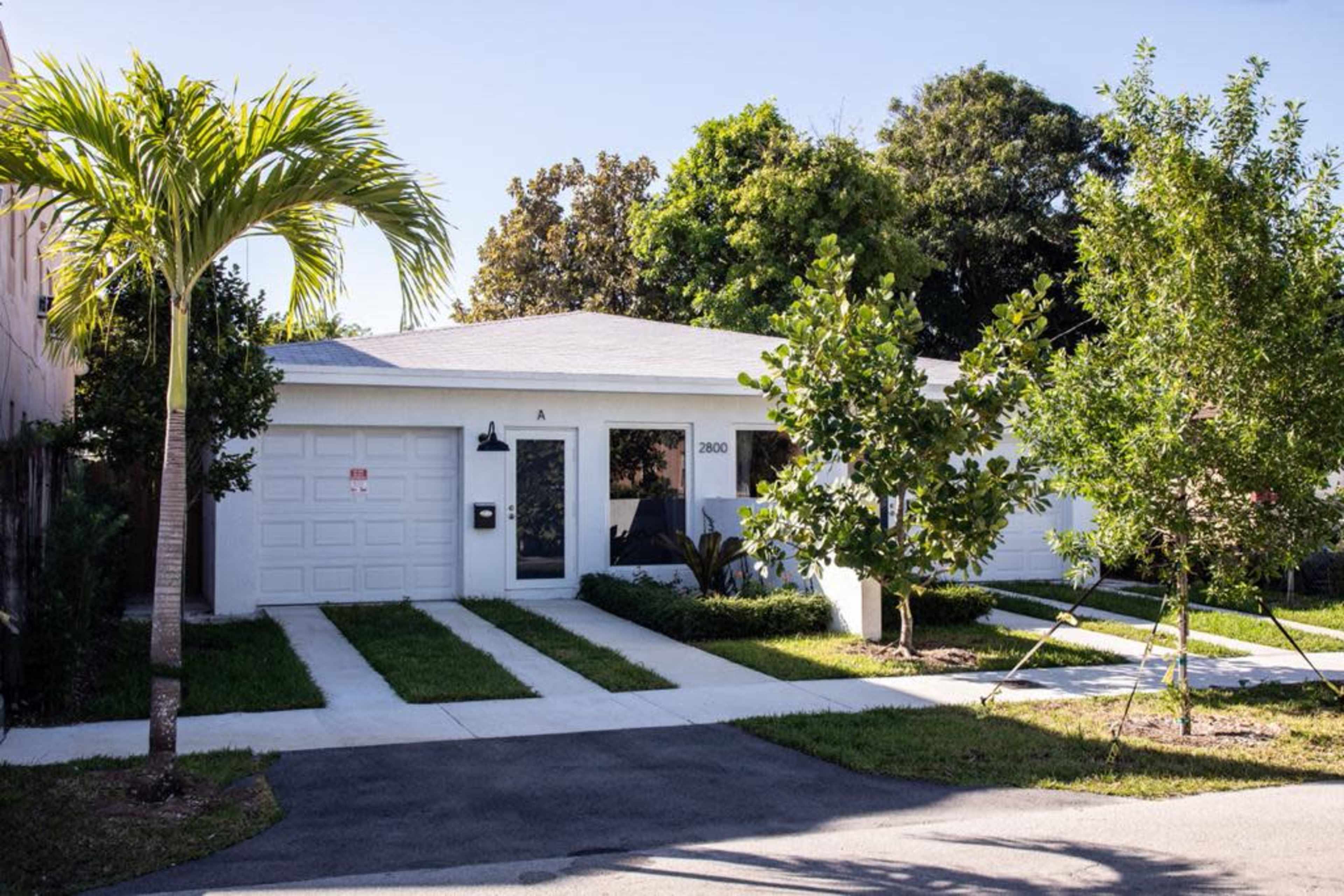 A single-story house with a garage, white exterior, and landscaped front yard is shown, featuring a driveway and palm trees.
