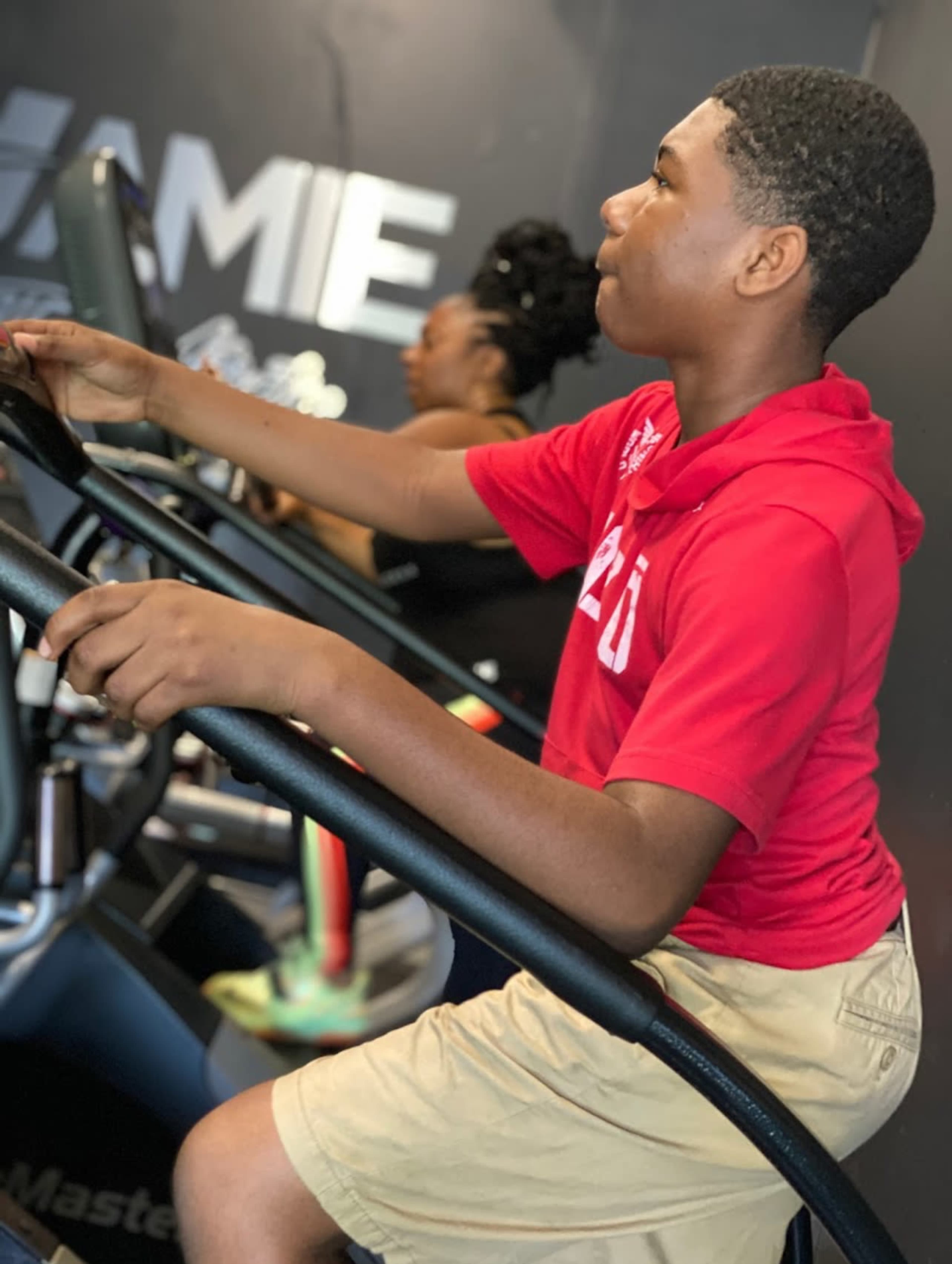 A young boy in a red hoodie exercises on a fitness machine while a woman workouts in the background.