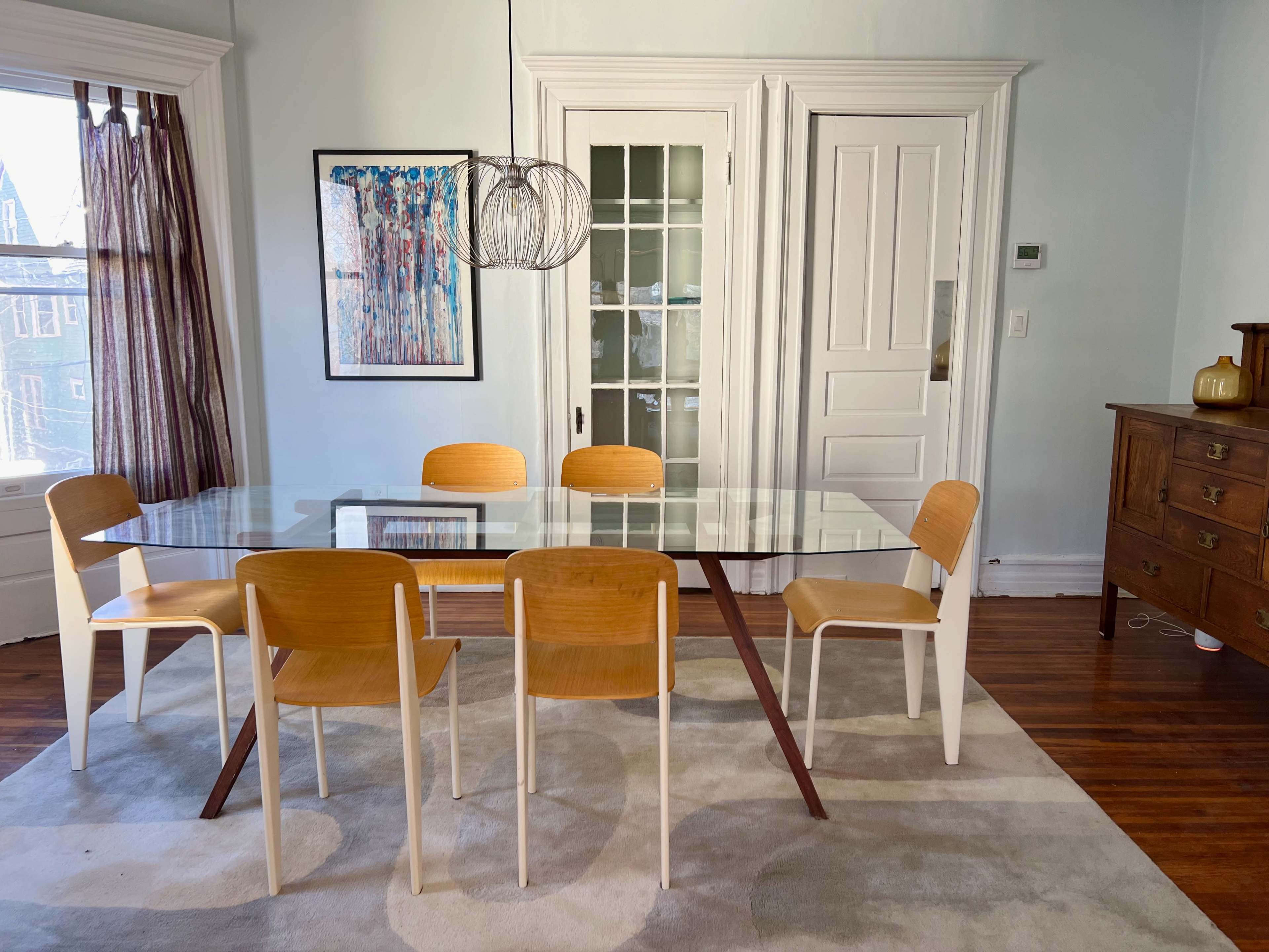 A modern dining room features a glass-top table surrounded by four wooden chairs, with a patterned artwork on the wall and a wooden dresser in the background.