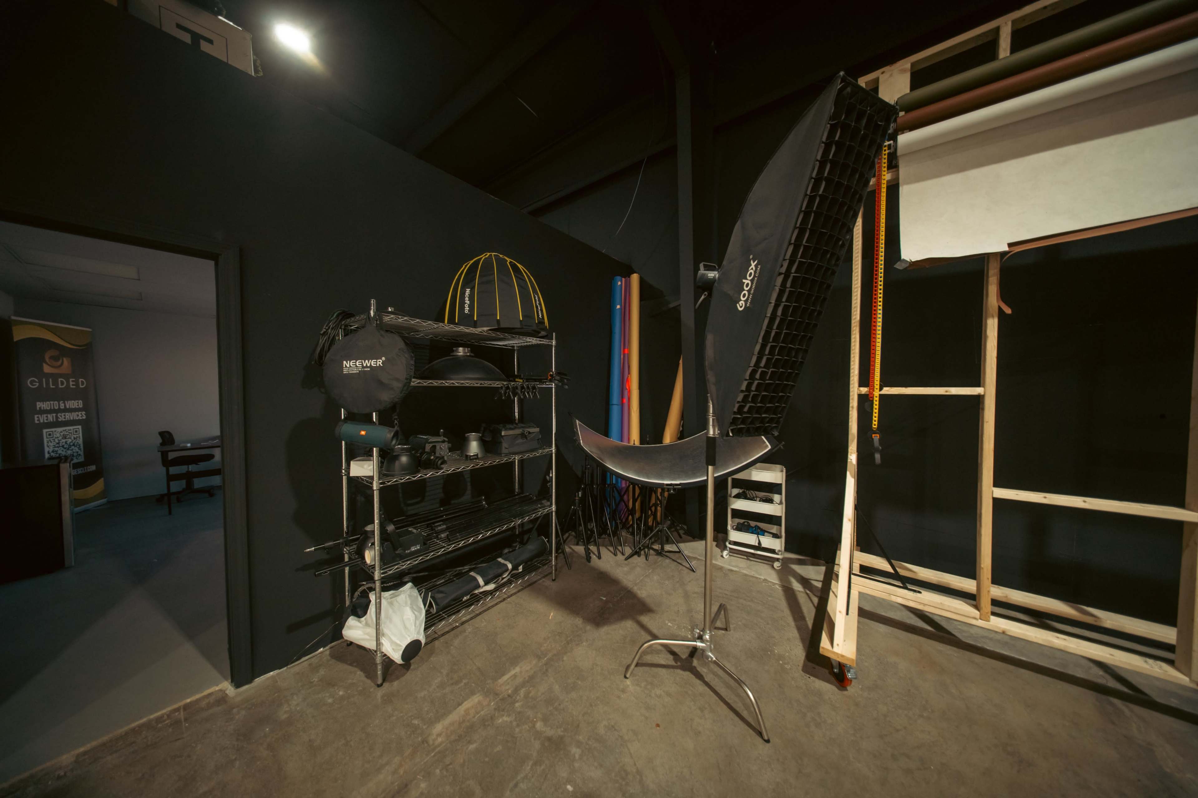 The image shows a photography studio with a black wall, a large softbox light on a stand, and a shelf filled with various photography equipment.