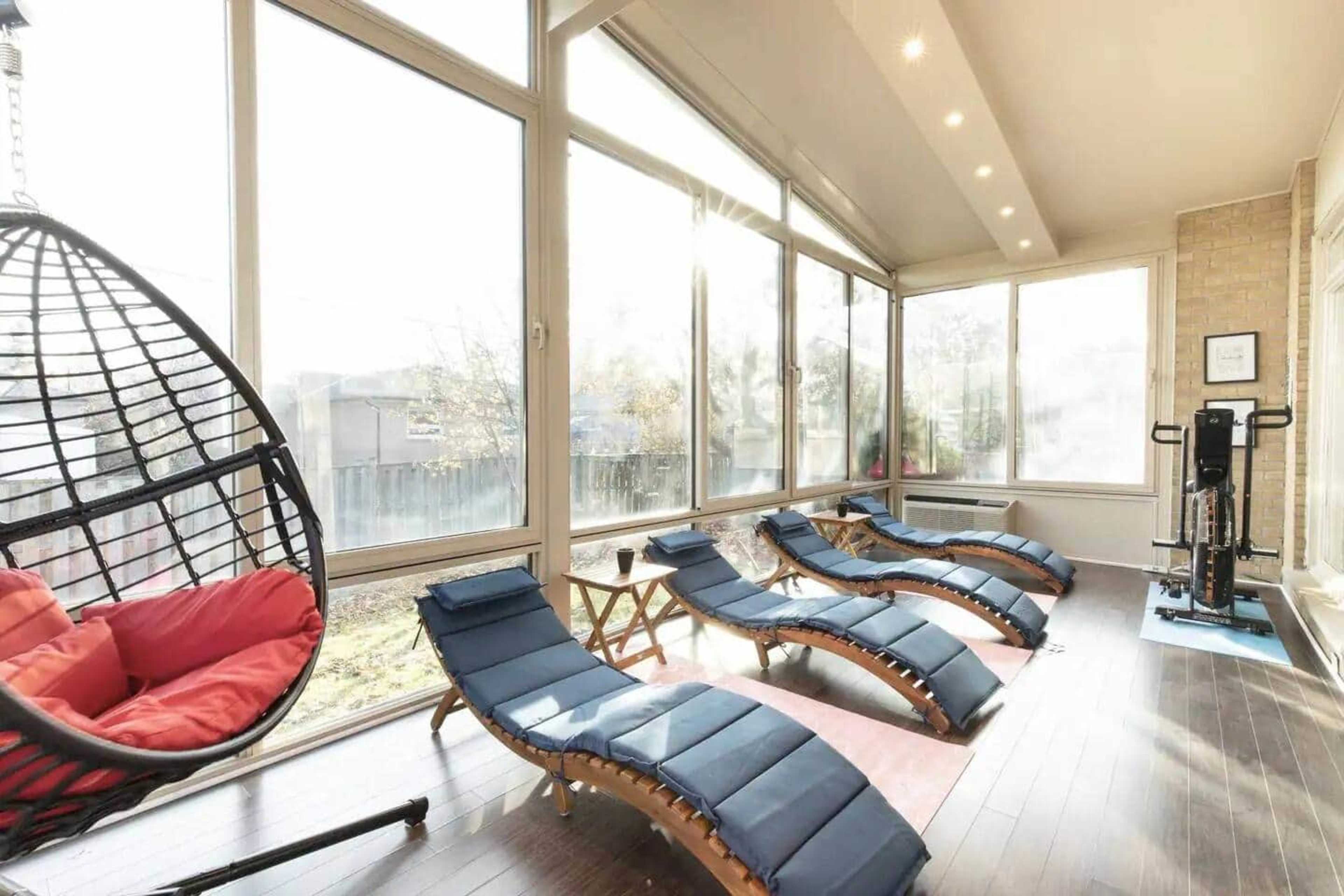 The image shows a sunlit room featuring several lounge chairs, a hanging chair, and exercise equipment, with large windows providing a view of the outdoors.
