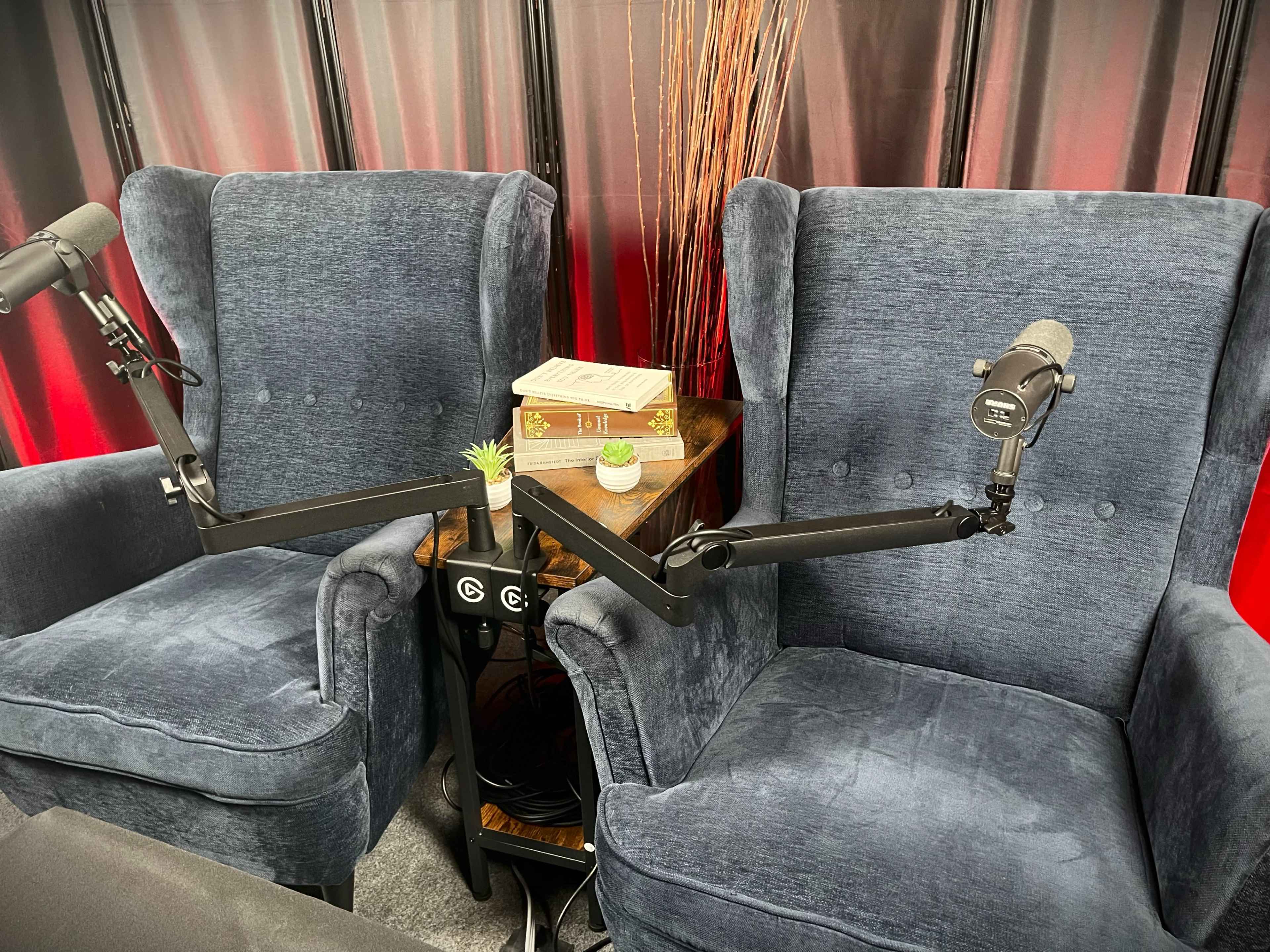 Two plush blue armchairs are positioned beside a small table with a stack of books and a decorative plant, each chair equipped with a microphone on an adjustable stand.