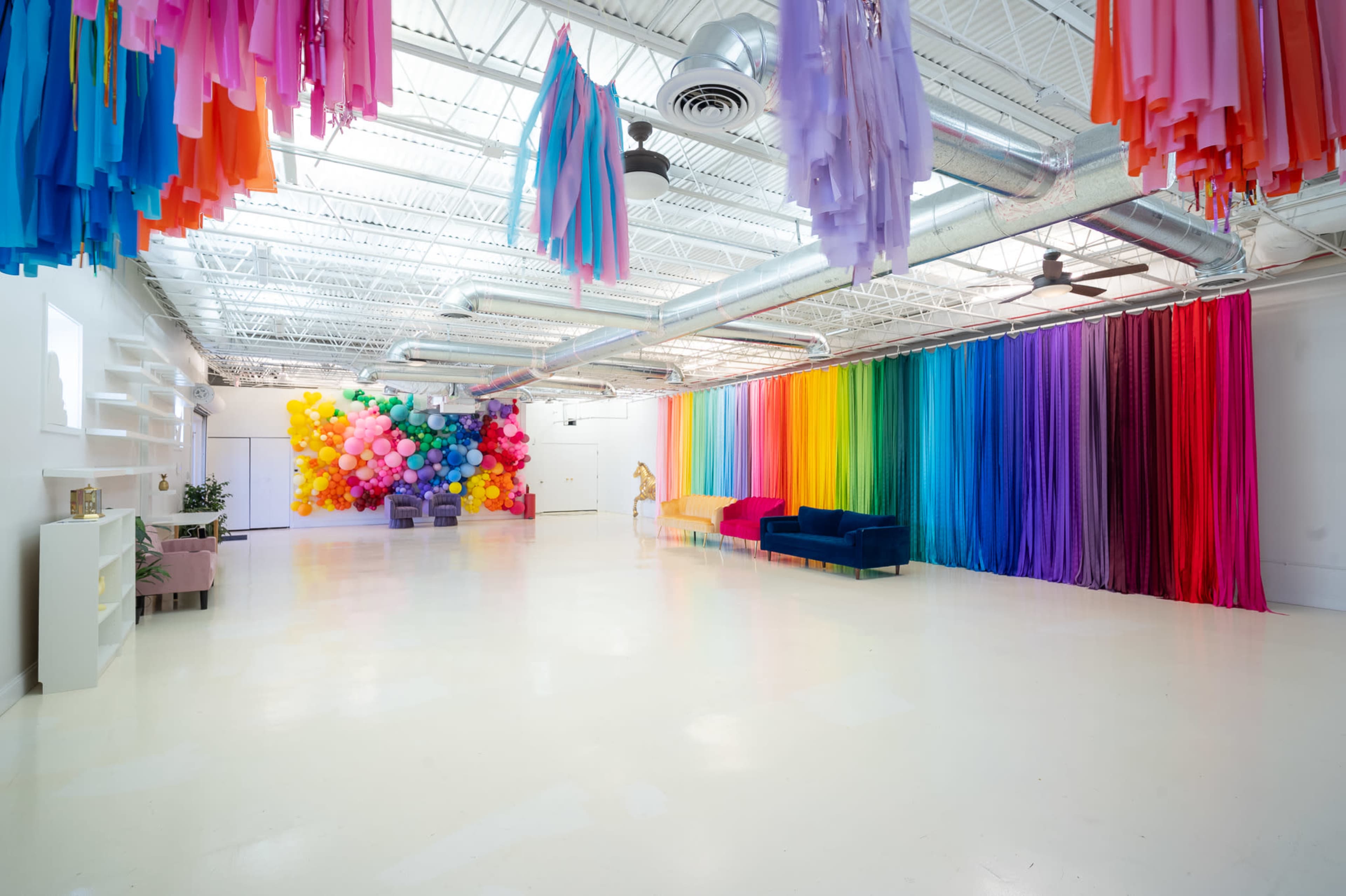 The image shows a spacious event room with colorful paper decorations hanging from the ceiling, a vibrant rainbow backdrop on one wall, and a cluster of balloons in various colors on the opposite side.