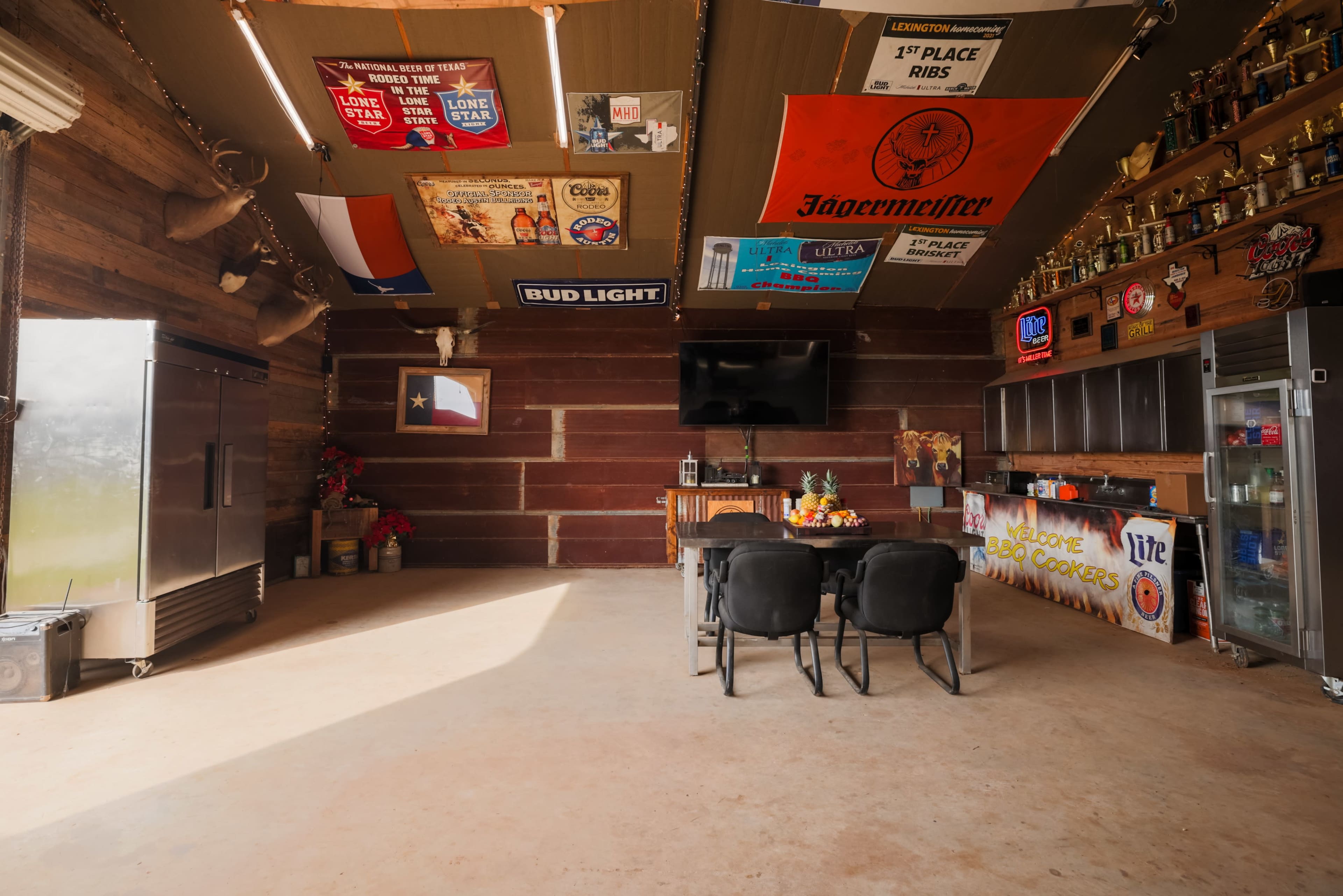 The image shows a rustic indoor space with a bar area, adorned with various banners and trophies, and equipped with a refrigerator and a television.