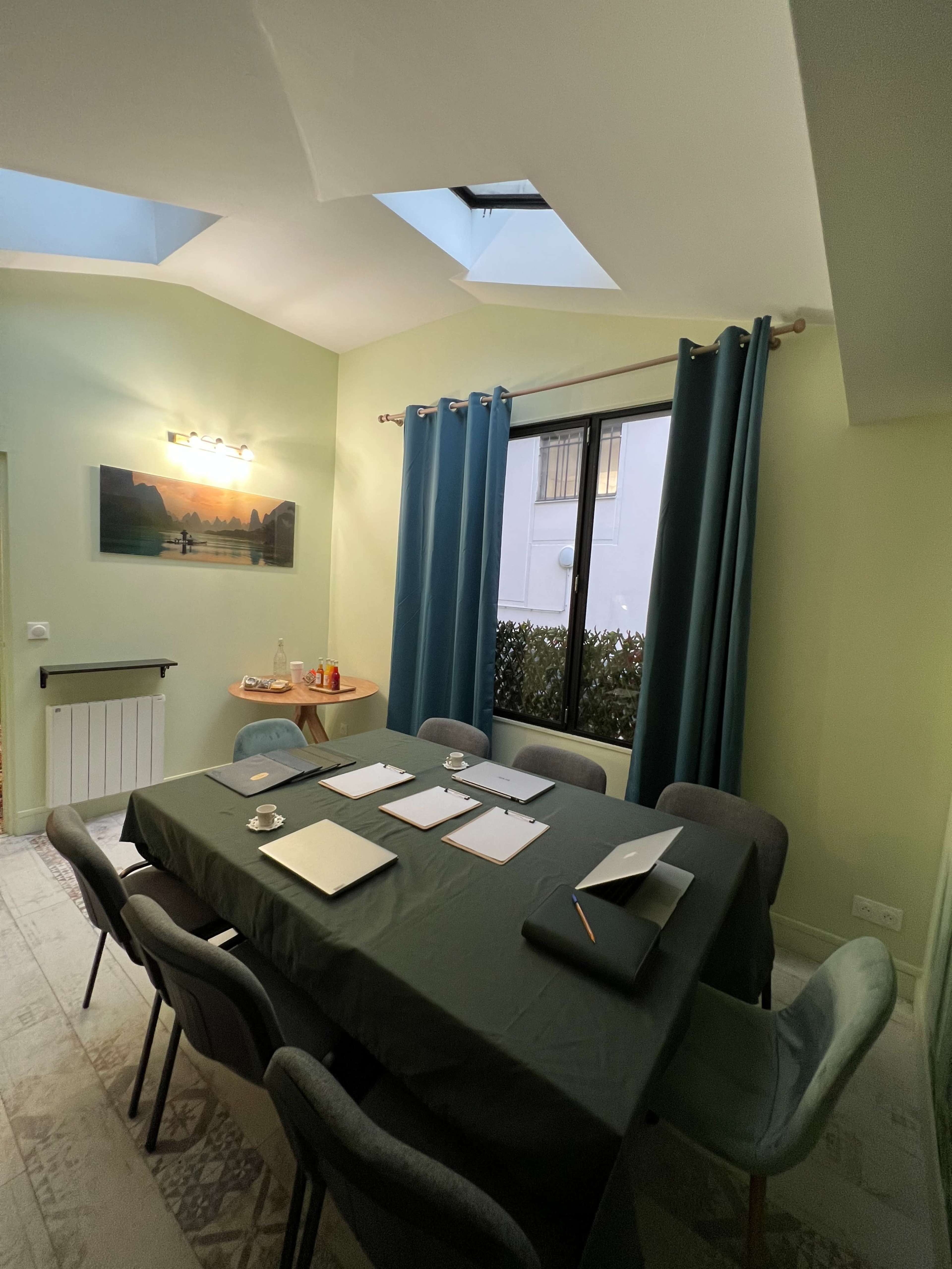A meeting room with a large green table covered with papers, a laptop, and a notepad, surrounded by chairs and illuminated by natural light from a skylight and a window.