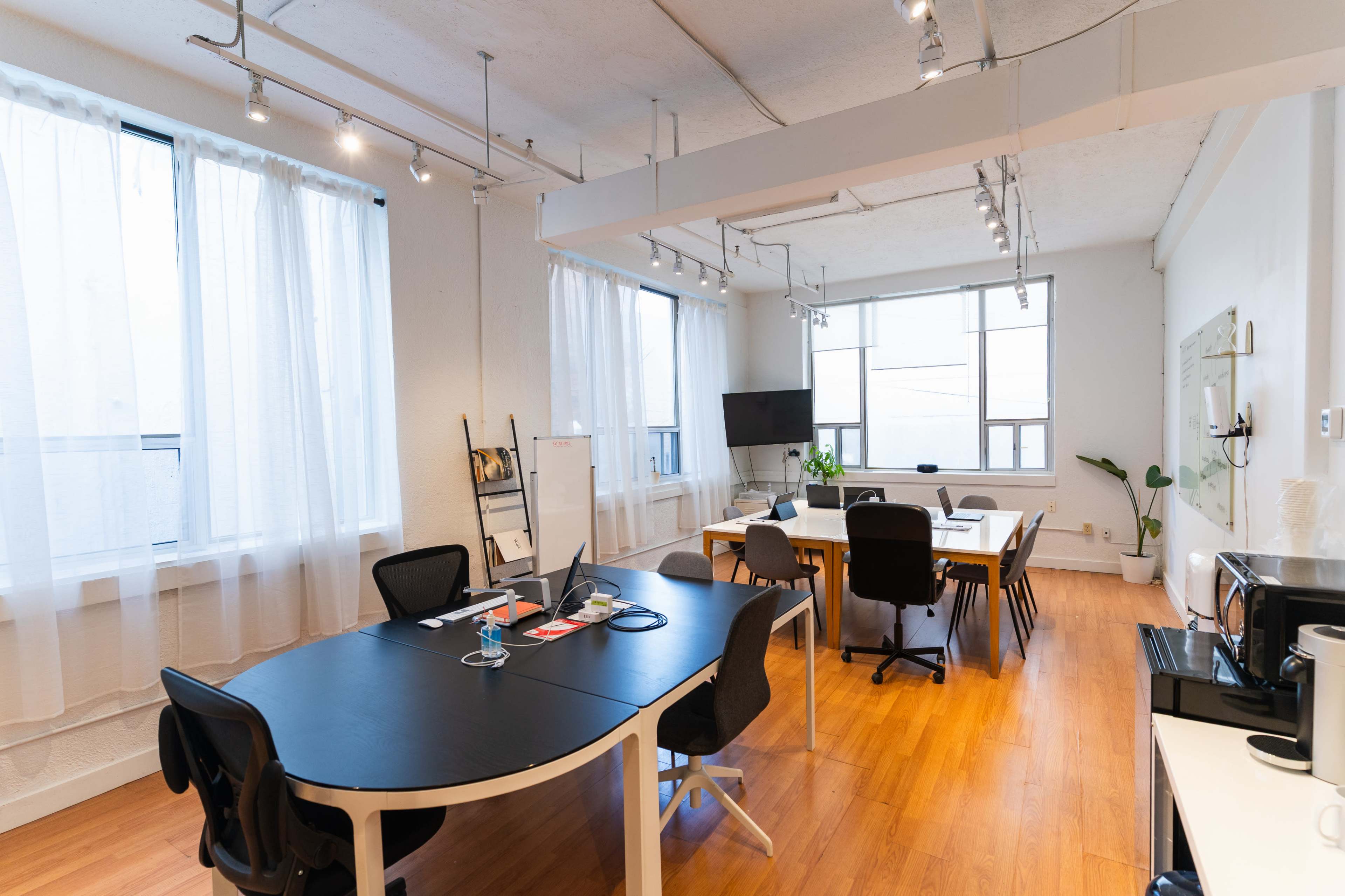 The image shows a modern office space with a large table, several chairs, and natural light streaming in through large windows.