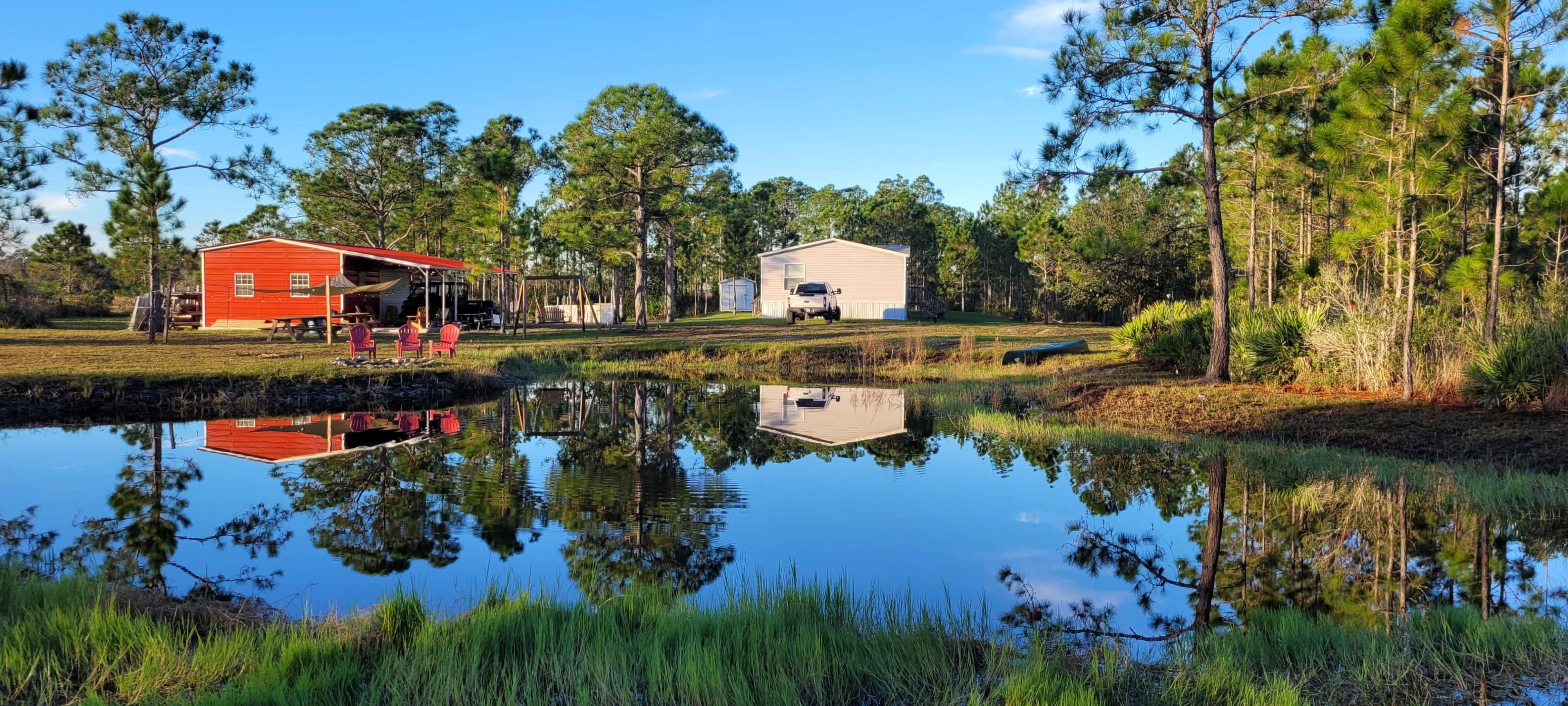 A peaceful scene shows two buildings by a pond, with trees and a clear sky reflecting in the water.