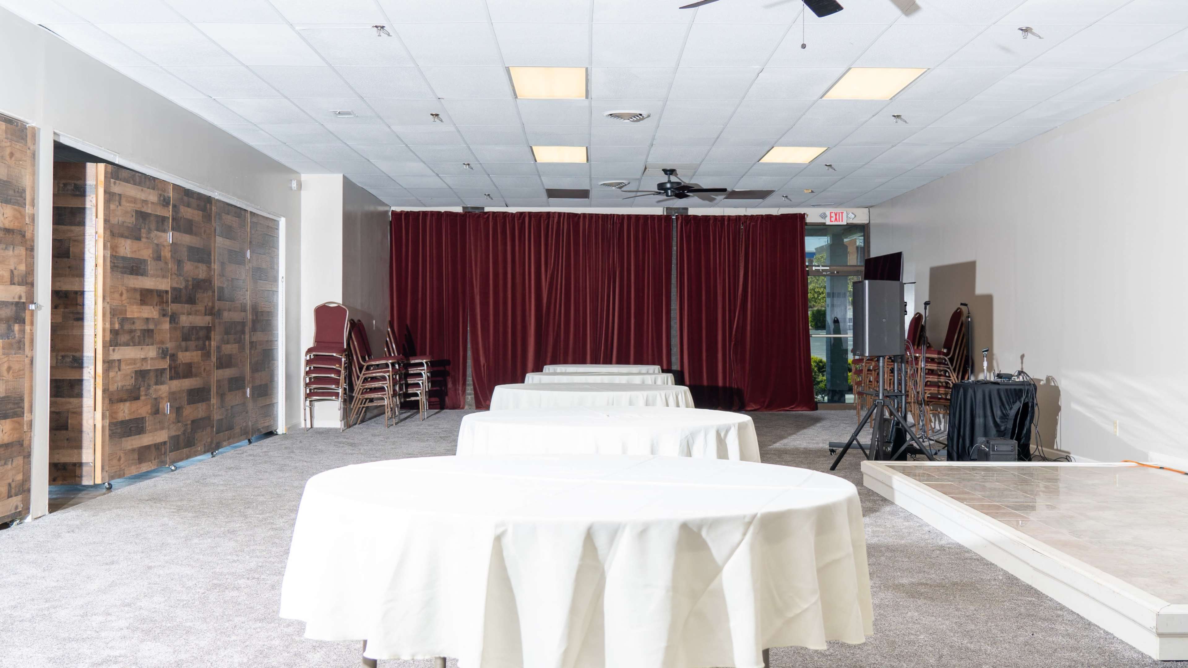 A banquet hall features several round tables covered with white tablecloths arranged in rows, facing a red curtain backdrop.