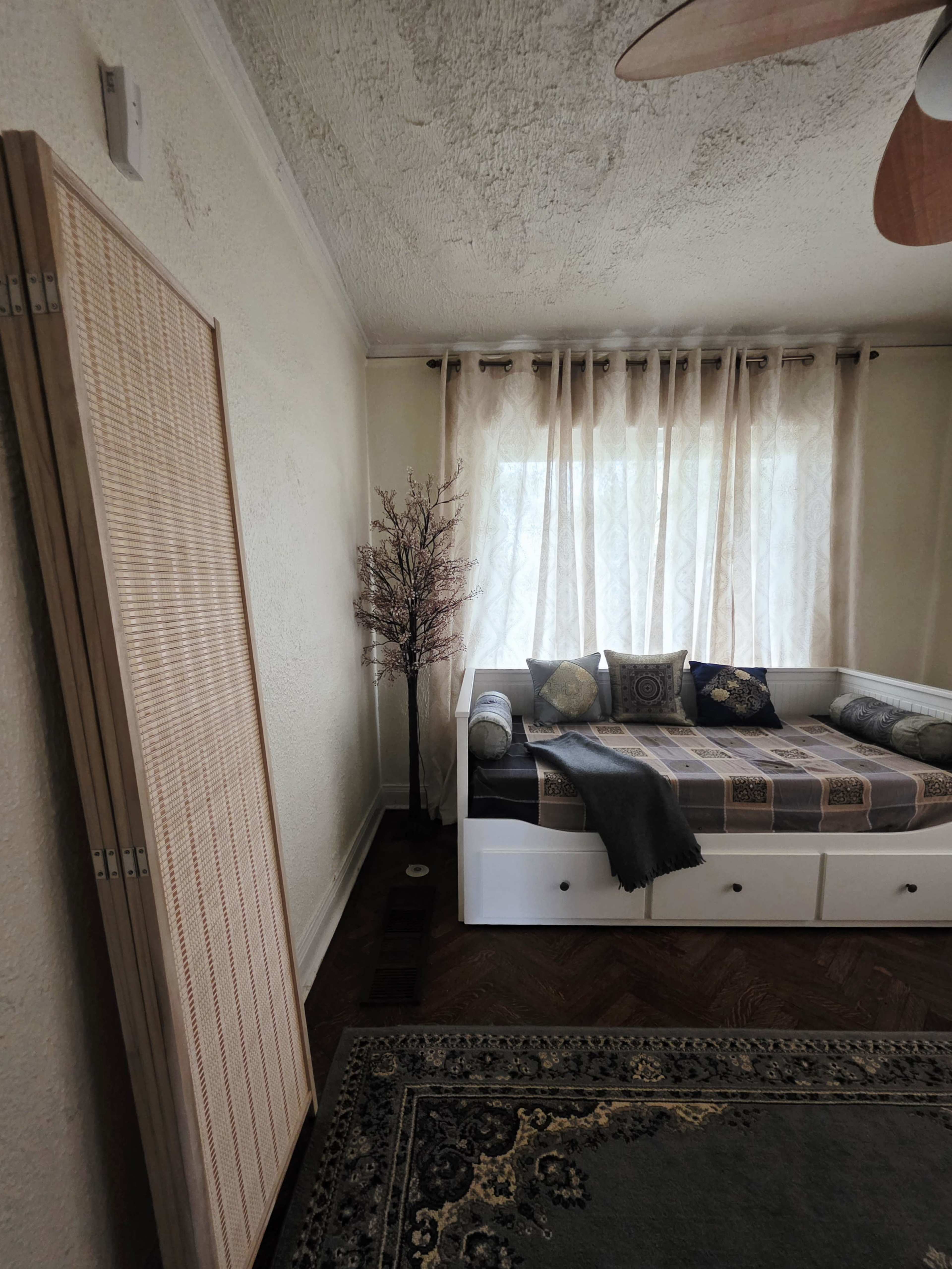 The image shows a bright room featuring a daybed with decorative pillows, a tall faux tree, a patterned area rug, and a wooden folding screen leaning against the wall.