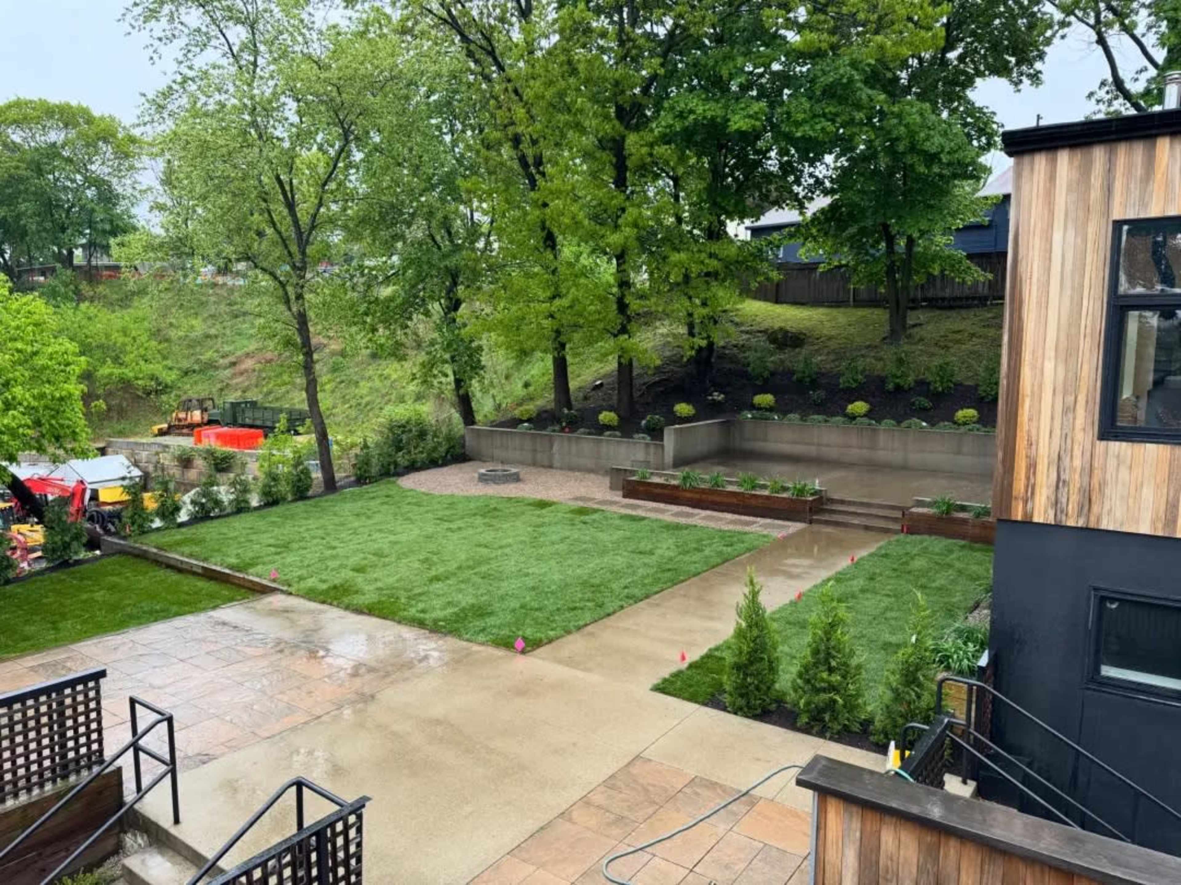 The image shows a landscaped yard with a newly installed lawn, surrounded by trees, stone pathways, and tiered planting areas.