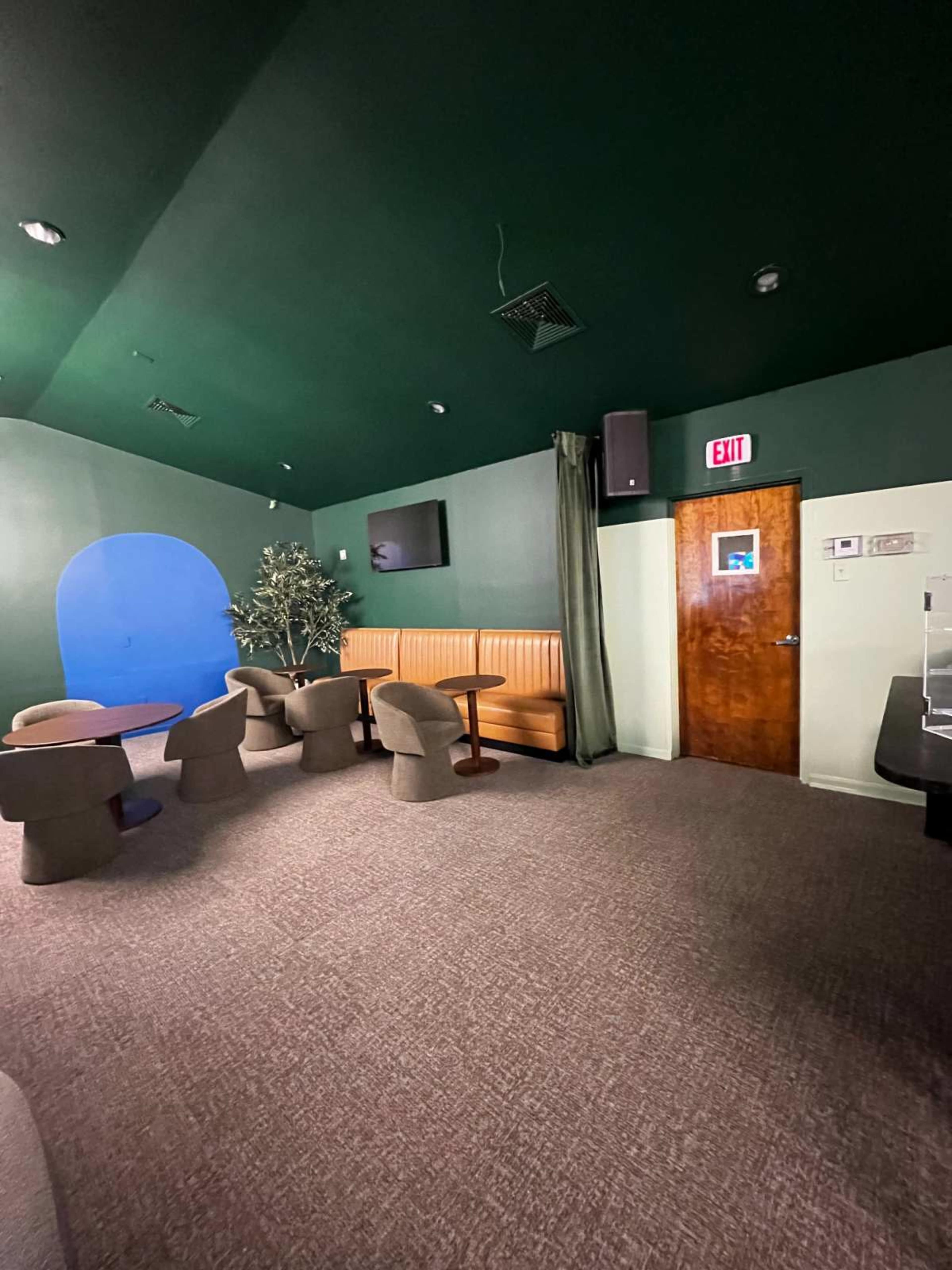 The image shows a room with green walls, featuring orange seating along one side, a door, and two small tables in a carpeted area.