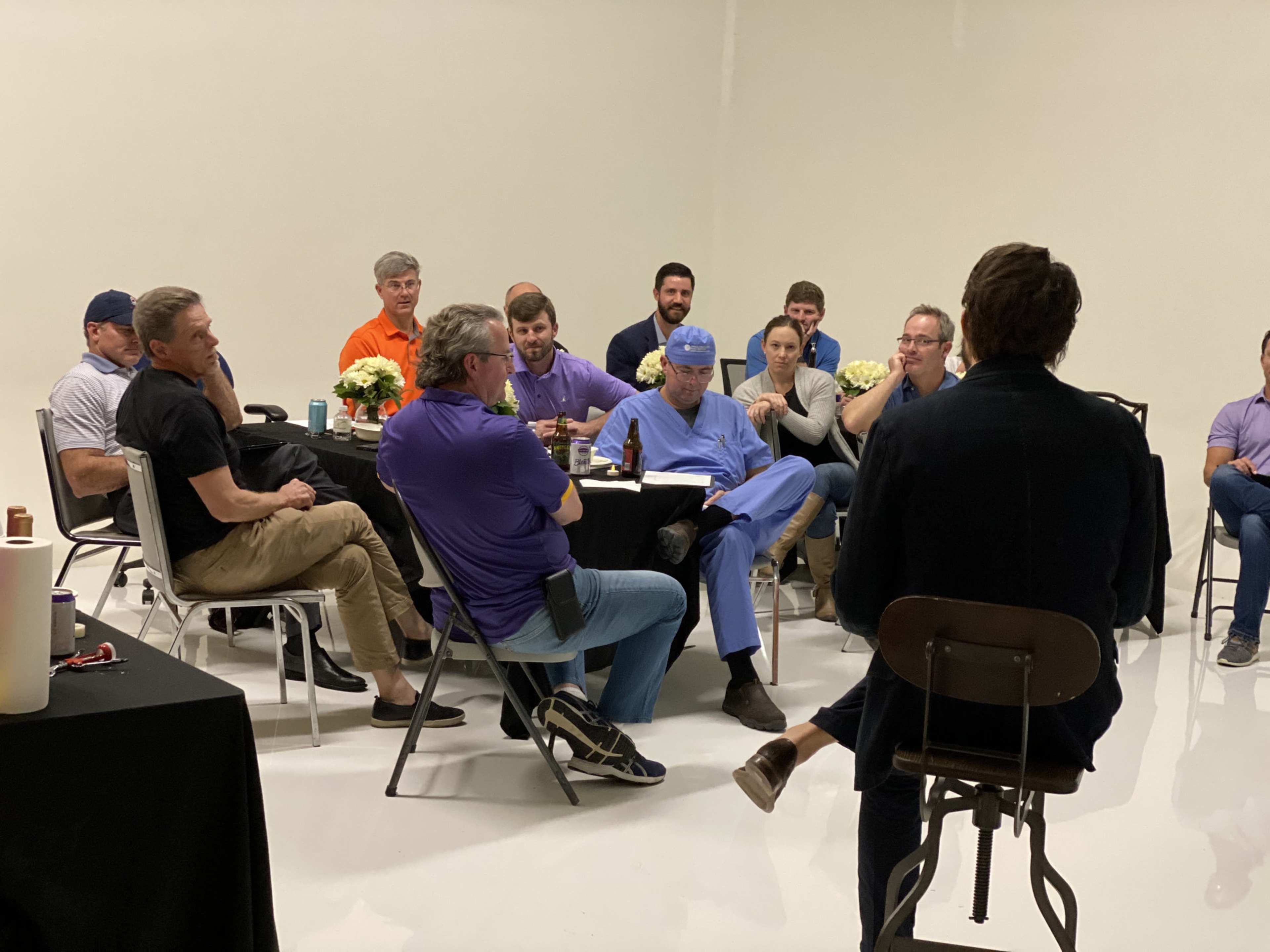A group of people, including individuals in surgical scrubs, sit around a table engaged in conversation in a well-lit room.