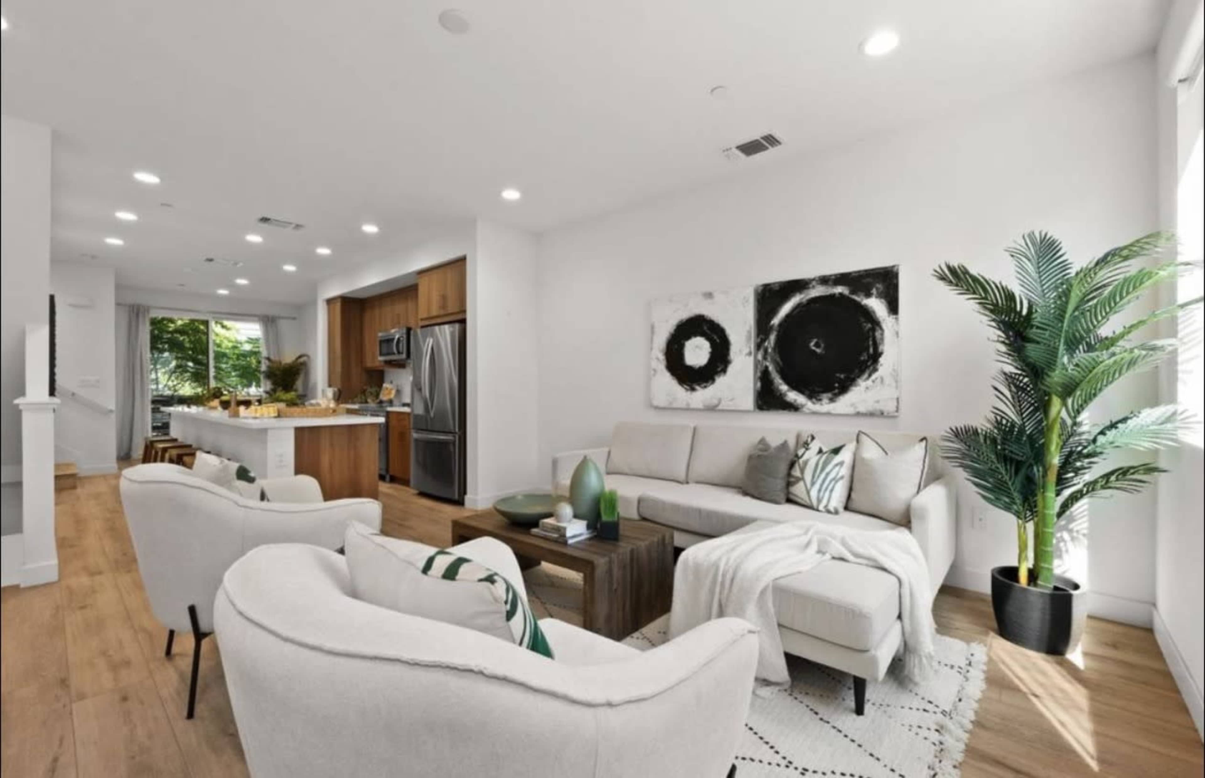 The image shows a modern living room with a light-colored sofa, two armchairs, and a coffee table, adjacent to a kitchen with wooden cabinets and stainless steel appliances.
