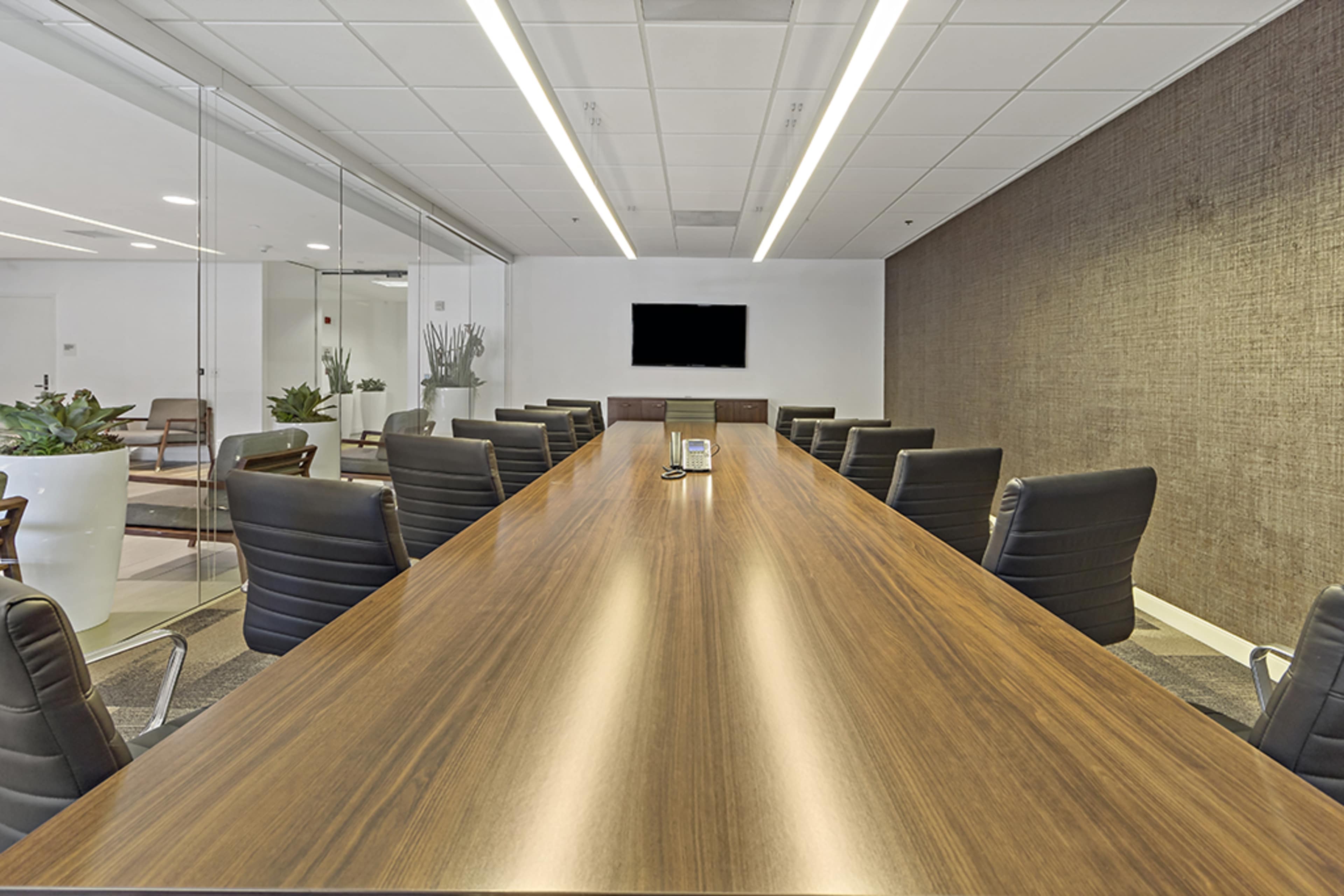 A modern conference room with a long wooden table, black leather chairs, and a wall-mounted television.