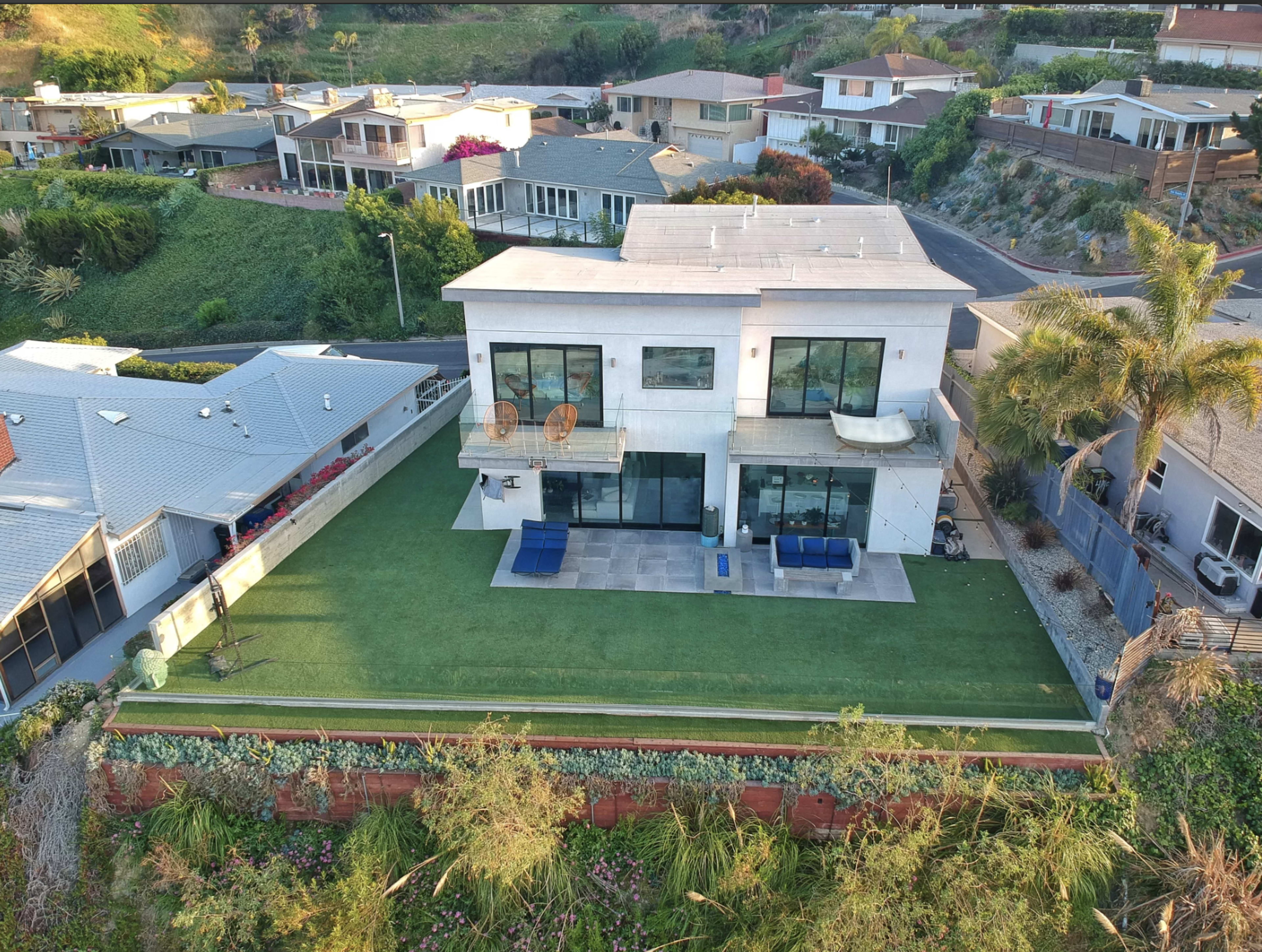 A modern two-story house with large windows and a backyard featuring artificial turf, surrounded by other residential homes on a hillside.