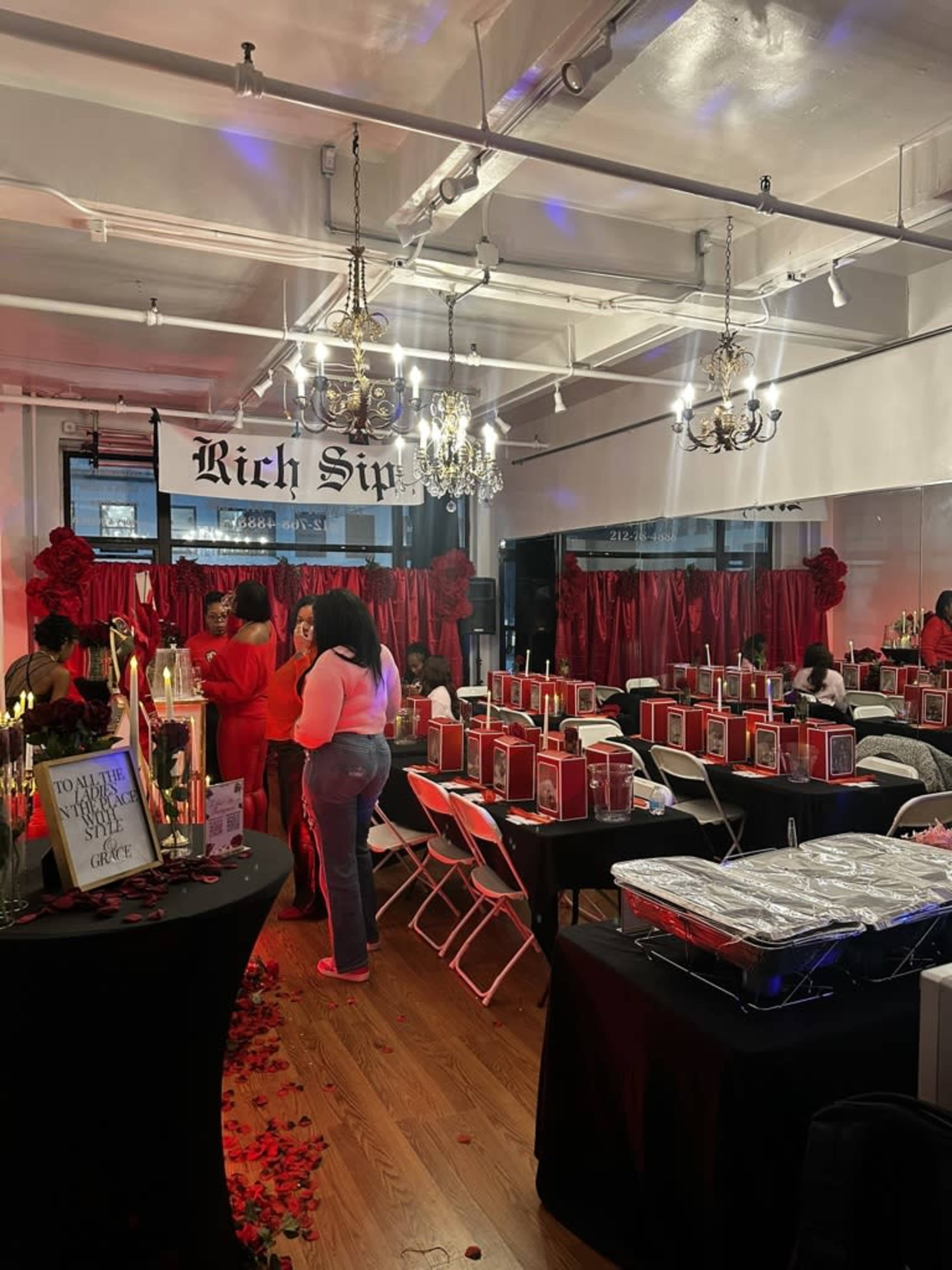 The image shows a decorated event space with tables set for a gathering, featuring red and black color schemes, and attendees mingling amidst floral accents and chandeliers.