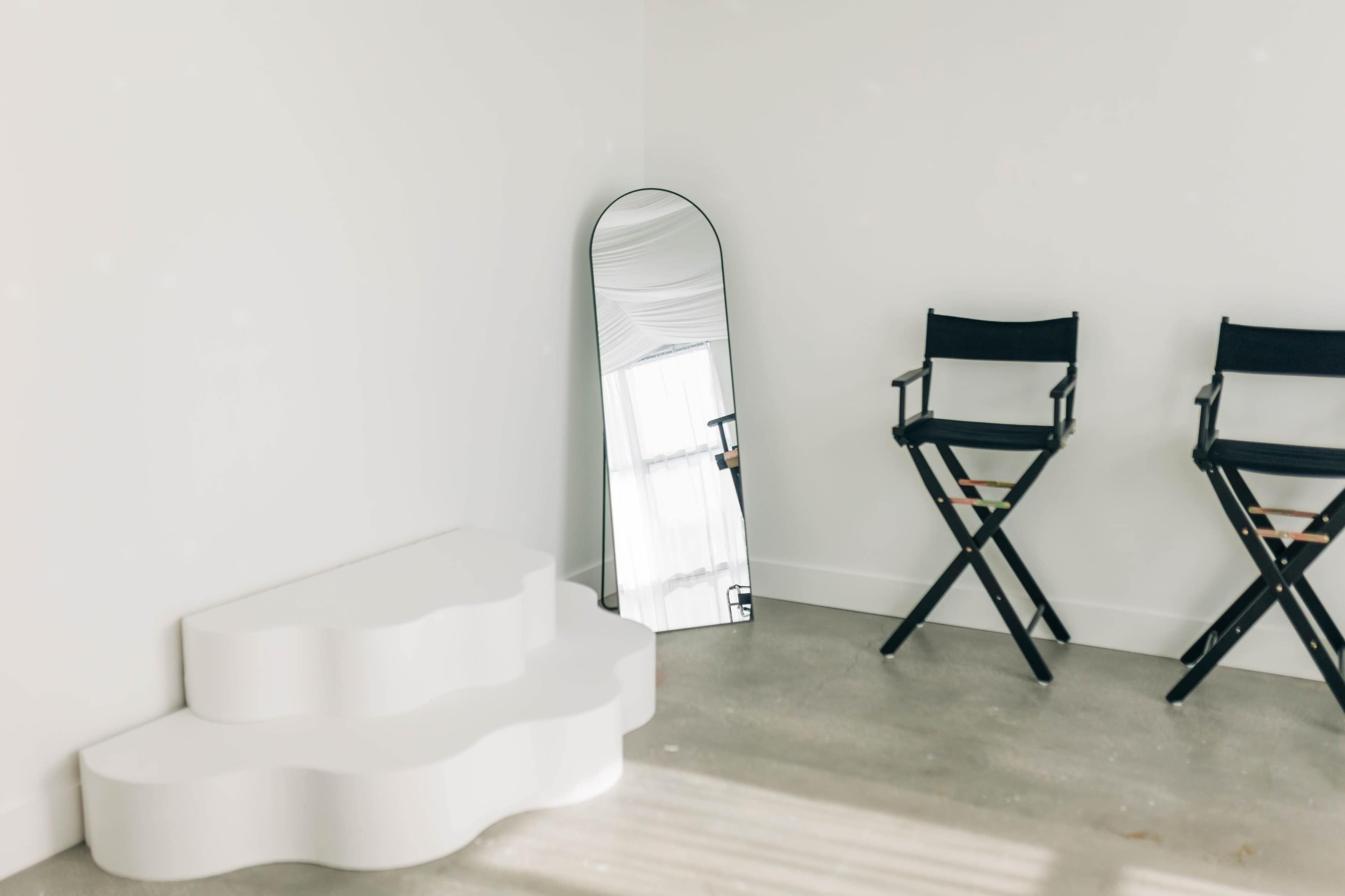 A minimalist room features a curved white platform, a tall mirror leaning against the wall, and a black director's chair.