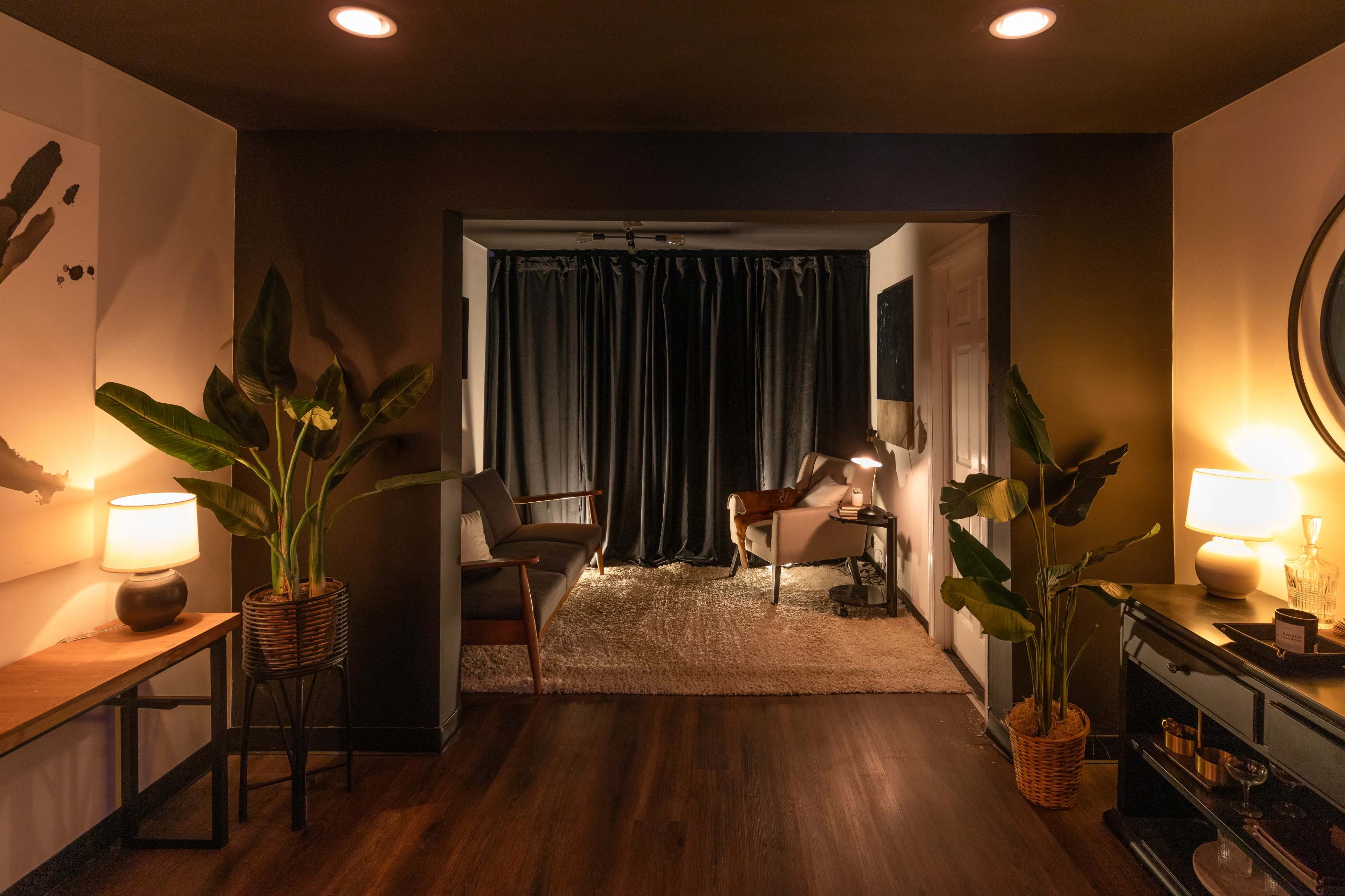 A cozy interior space features a dark room with a seating area, surrounded by plants, and illuminated by soft lighting.