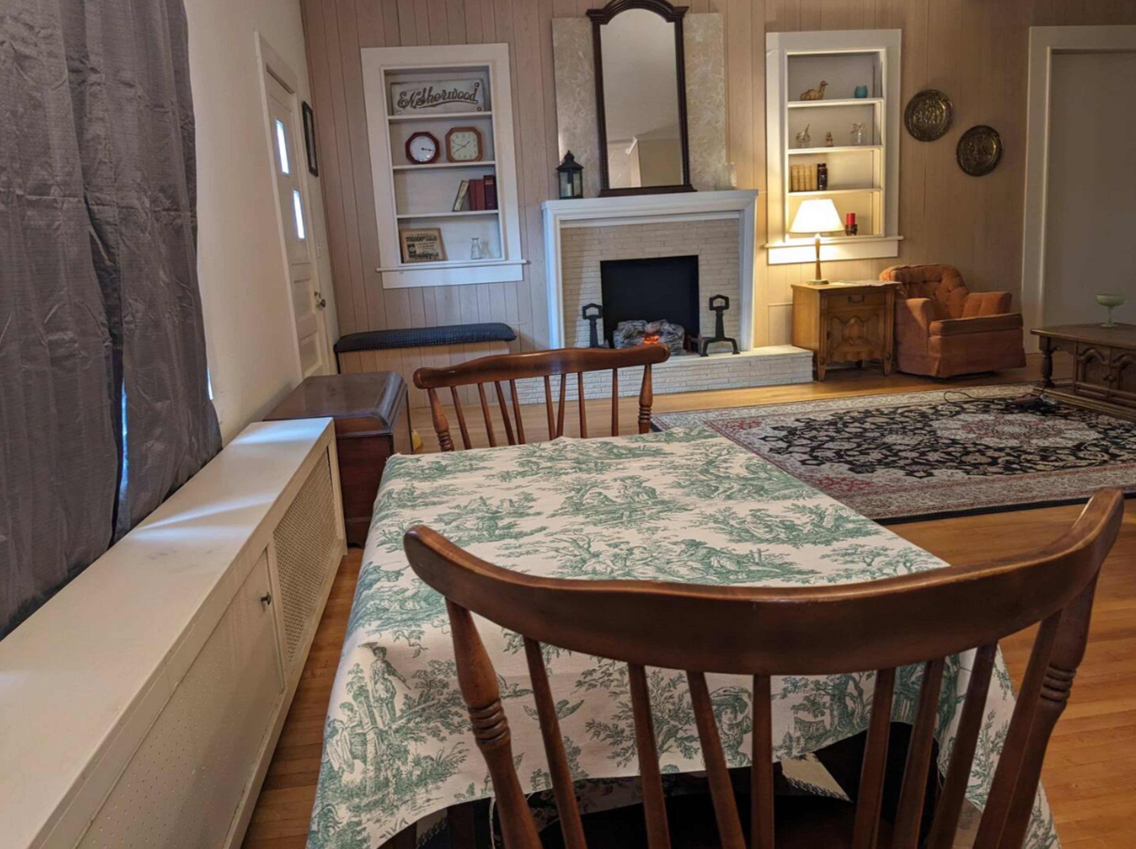 The image shows a cozy living room with a dining table in the foreground, a fireplace with shelves, and a seating area featuring a couch and armchair.